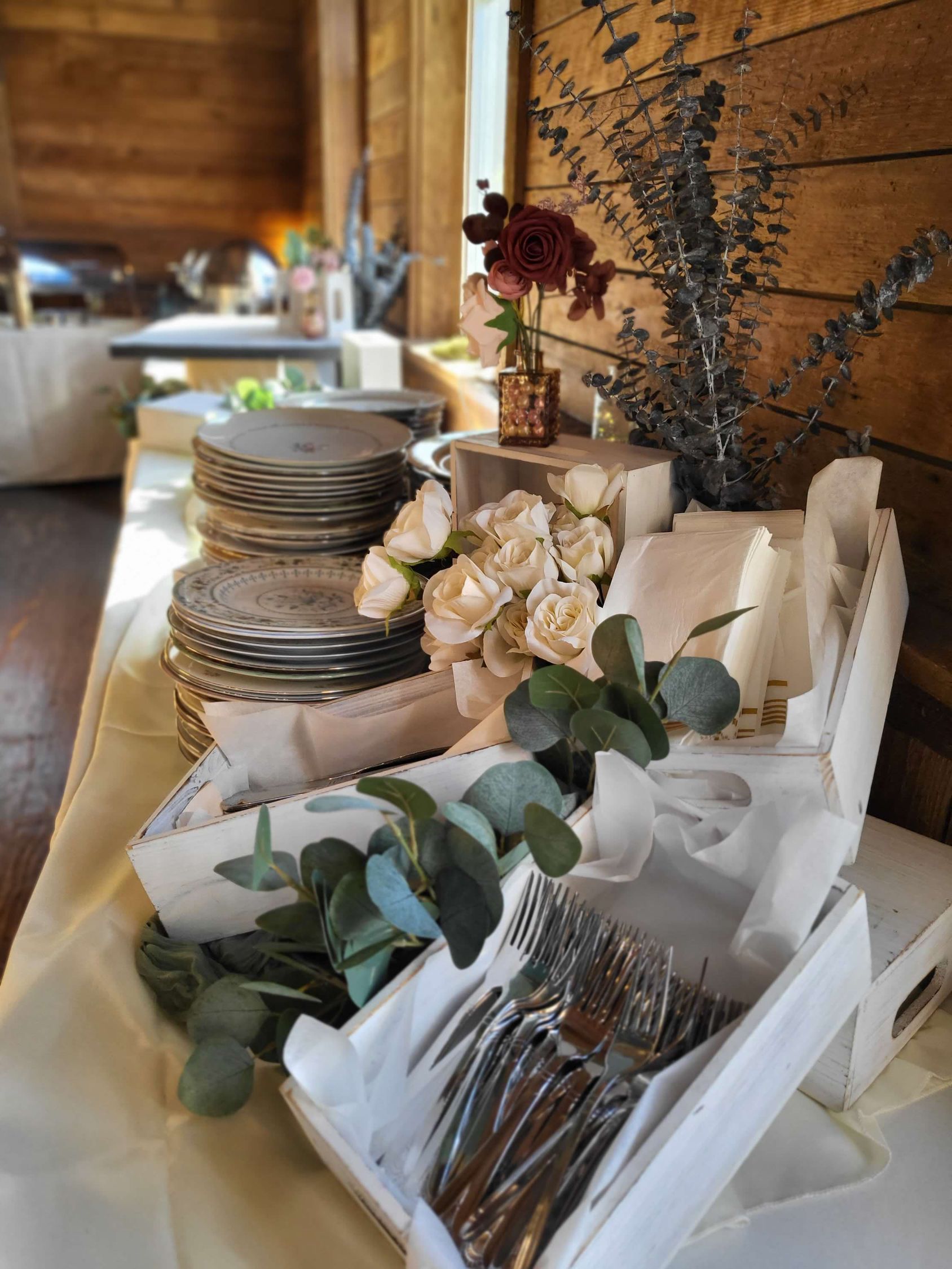 Buffet table with stacked plates, cutlery in a white box, and floral decorations. Wooden wall in the background.