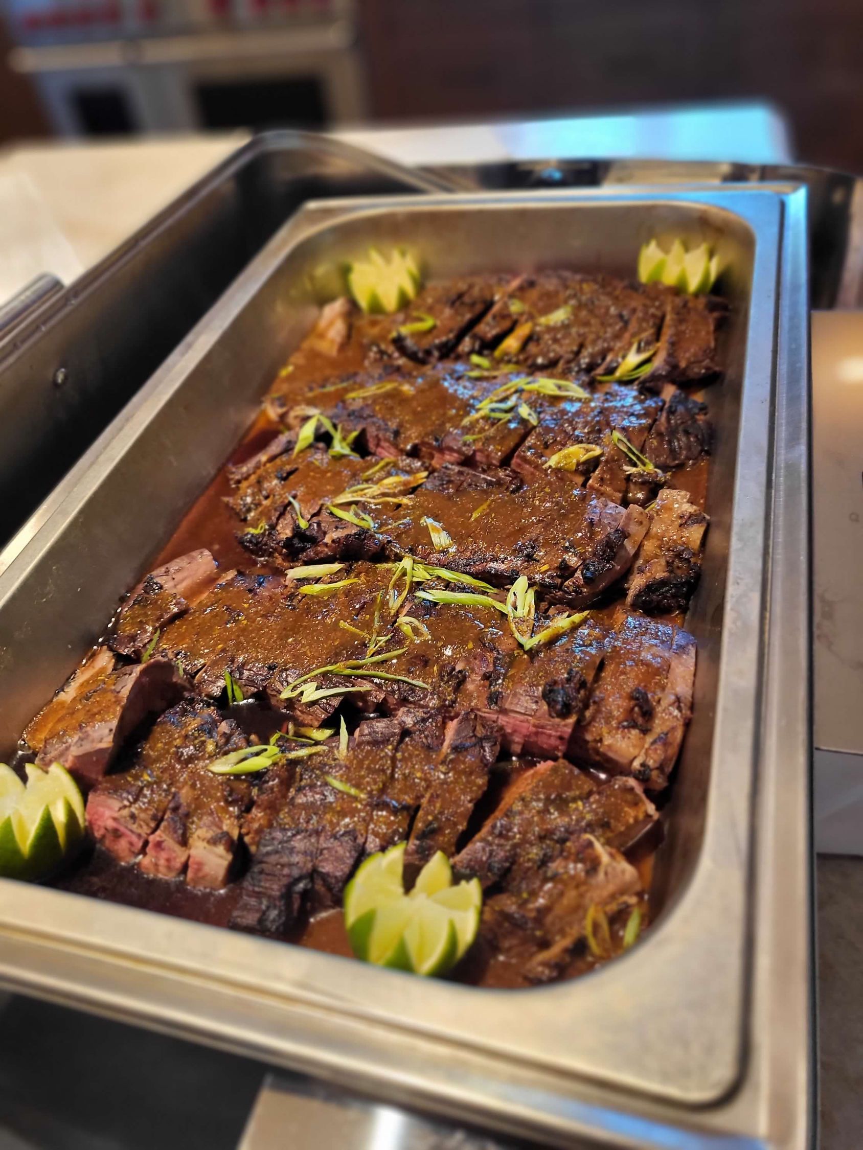 Beef brisket in a buffet tray, sliced and topped with sauce, garnished with lime.
