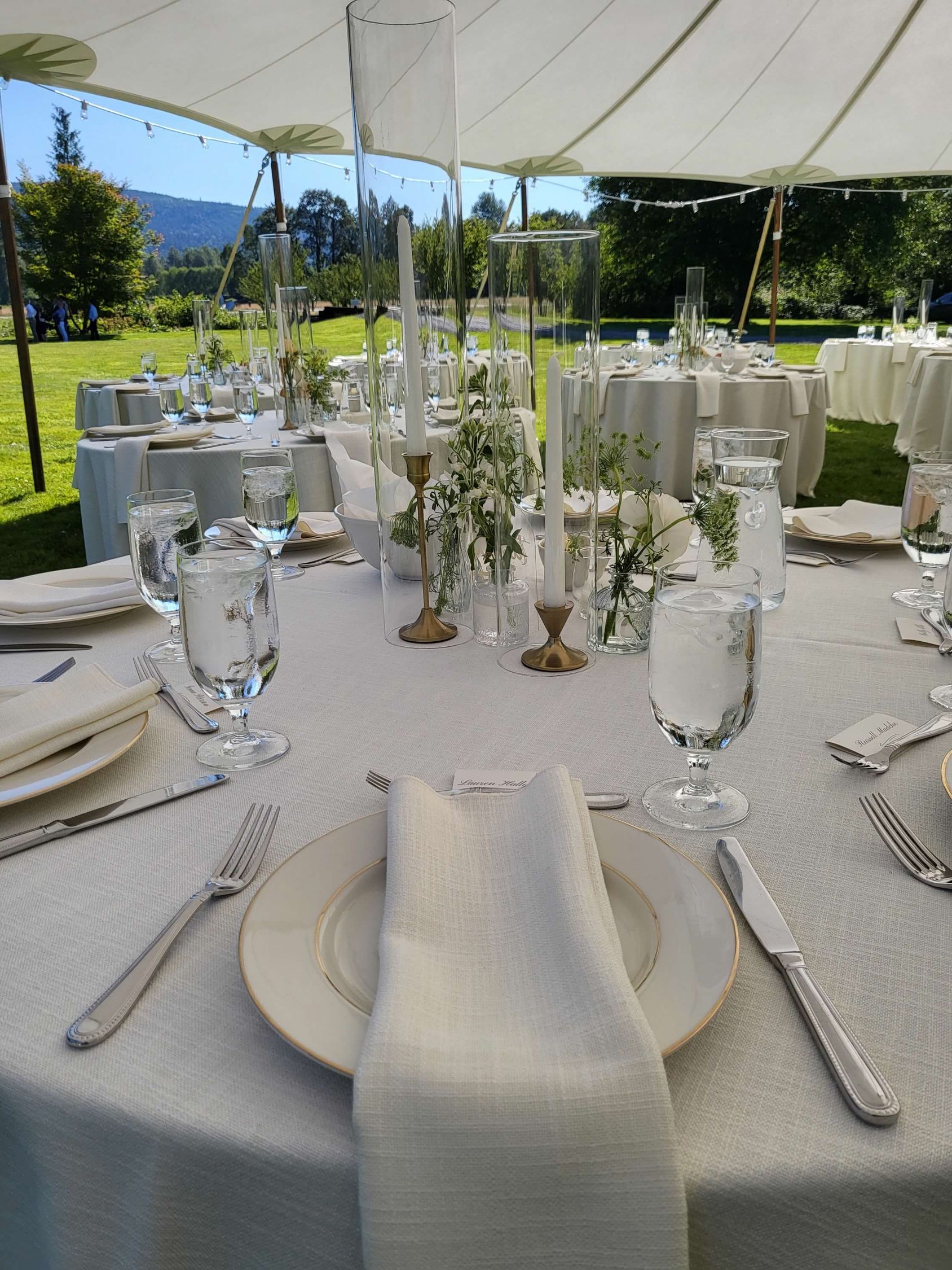 Outdoor reception table, white linen, glassware, floral centerpieces under a tent.
