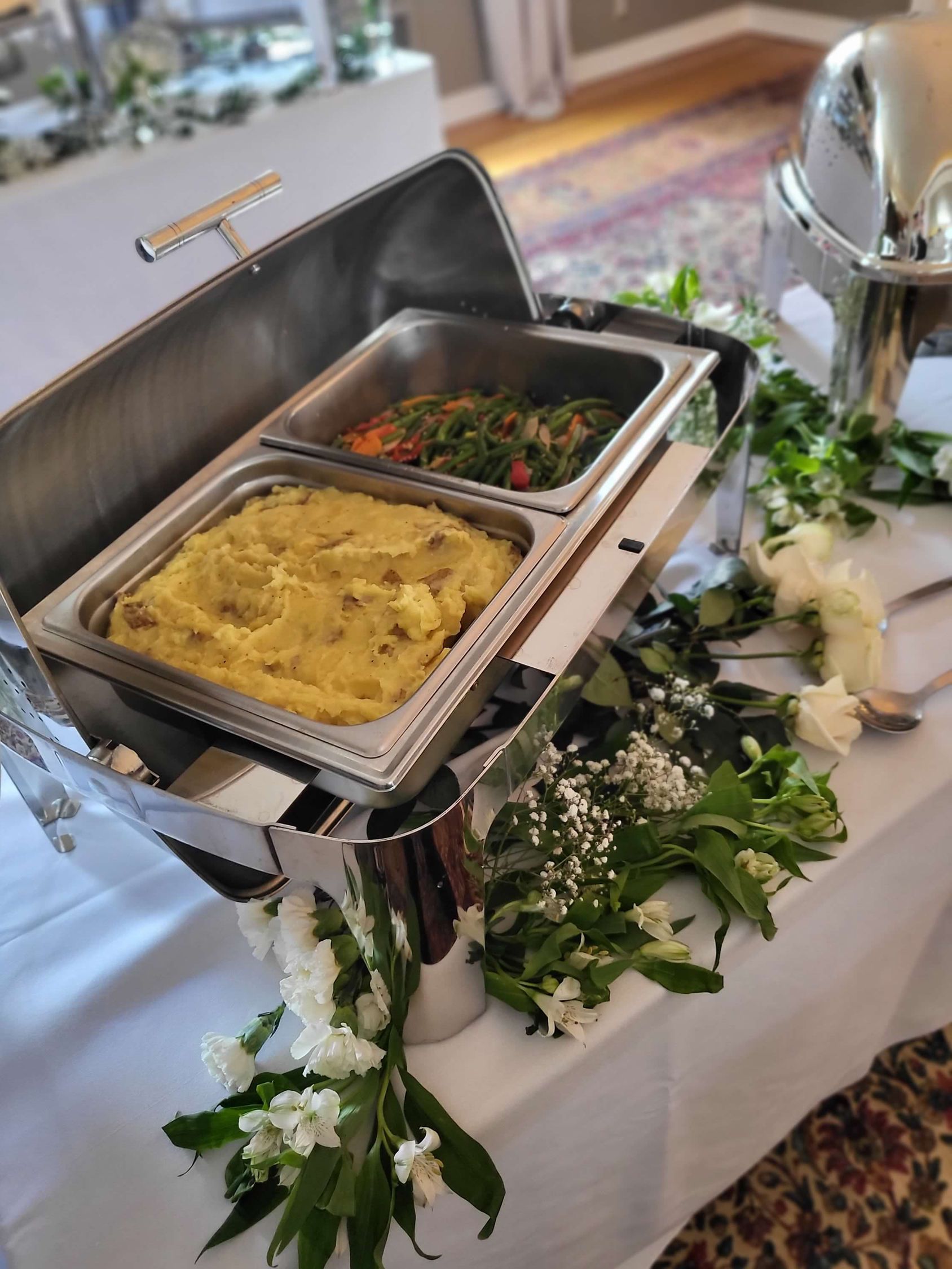 Silver chafing dish on a white table with two food trays, decorated with white flowers and greenery.