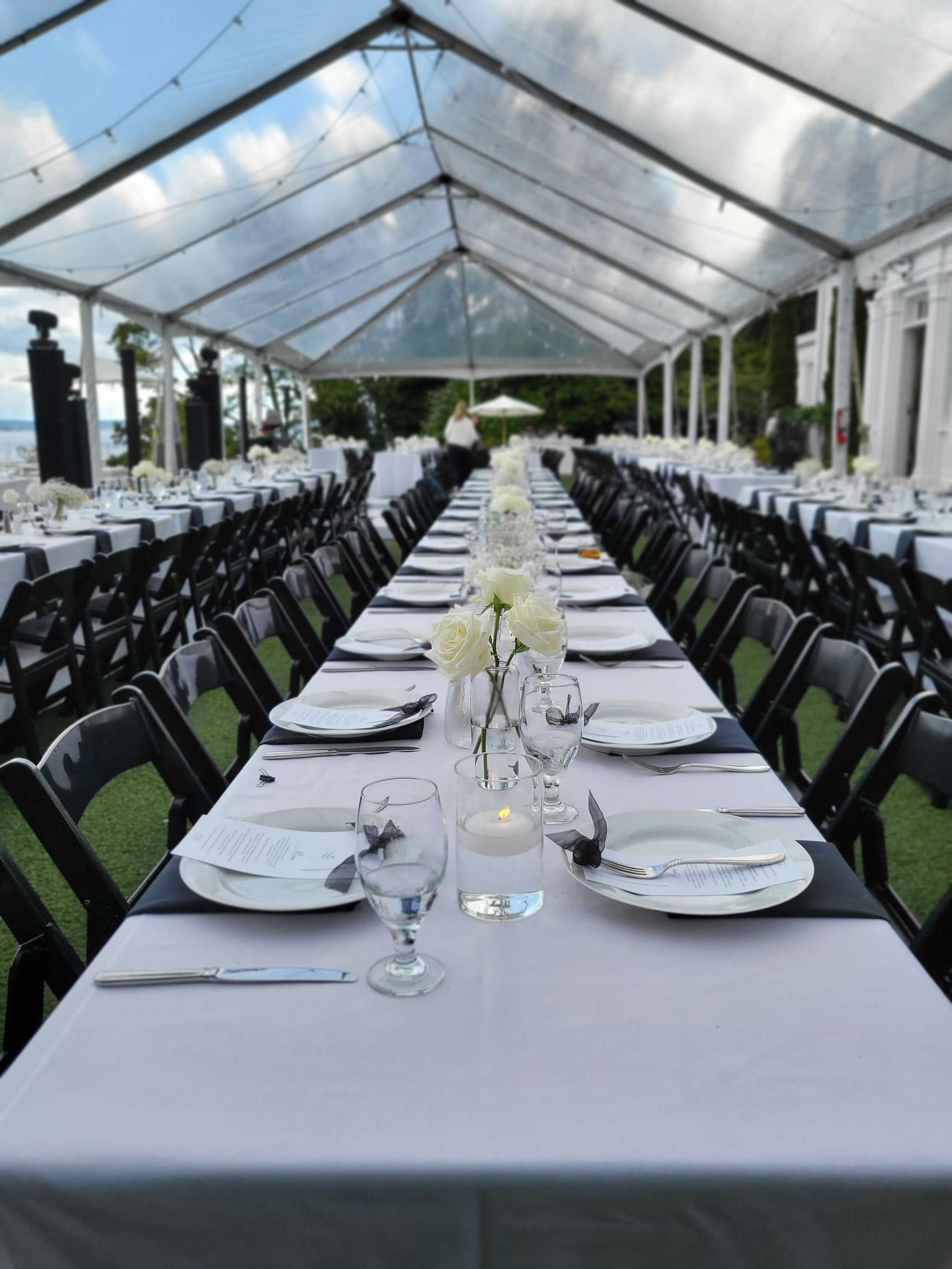 Long tables set for a formal event under a clear tent with black chairs, white linens, and floral centerpieces.
