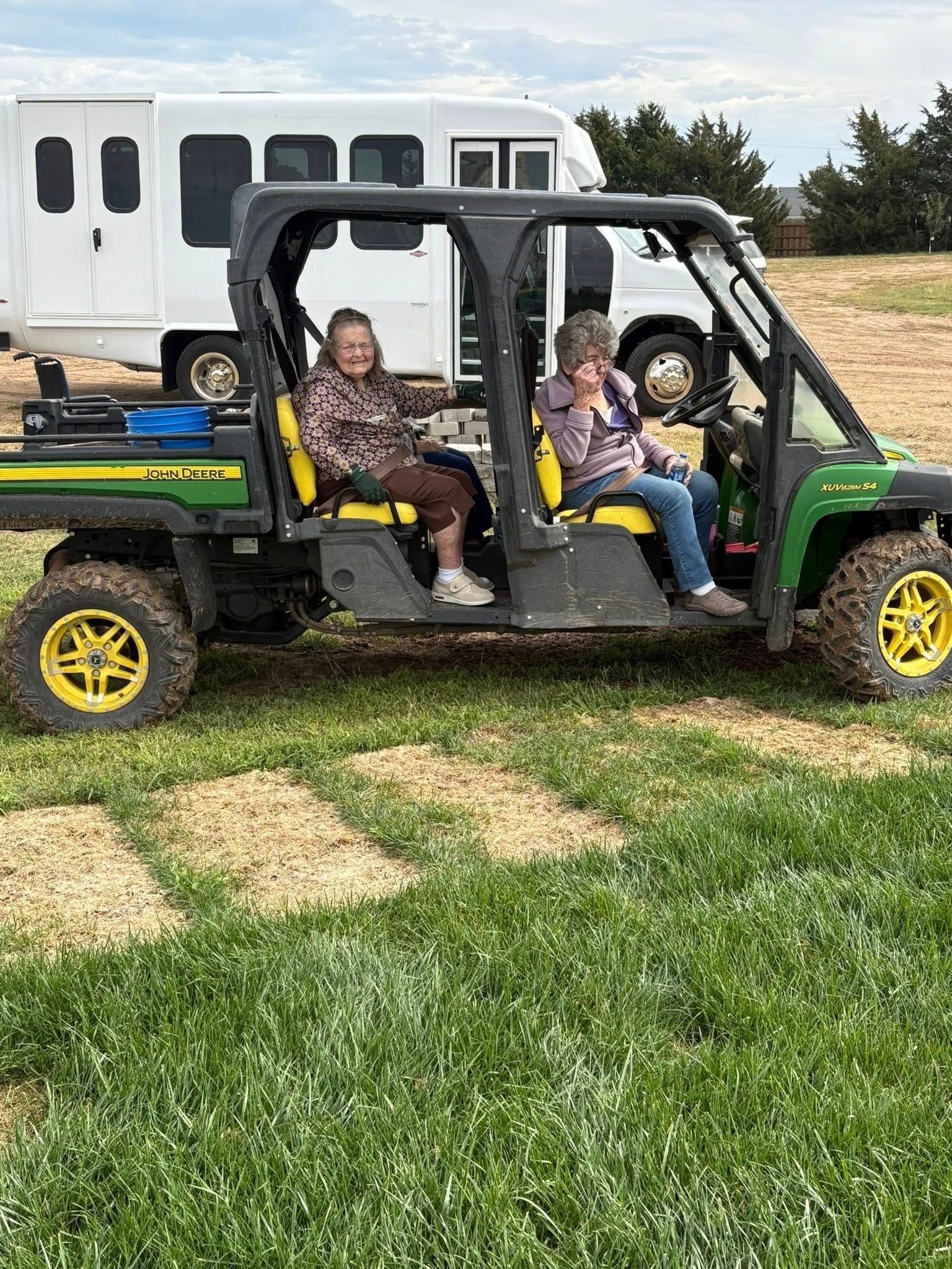 Two people sitting in a green John Deere utility vehicle in a field, with a white vehicle behind them.