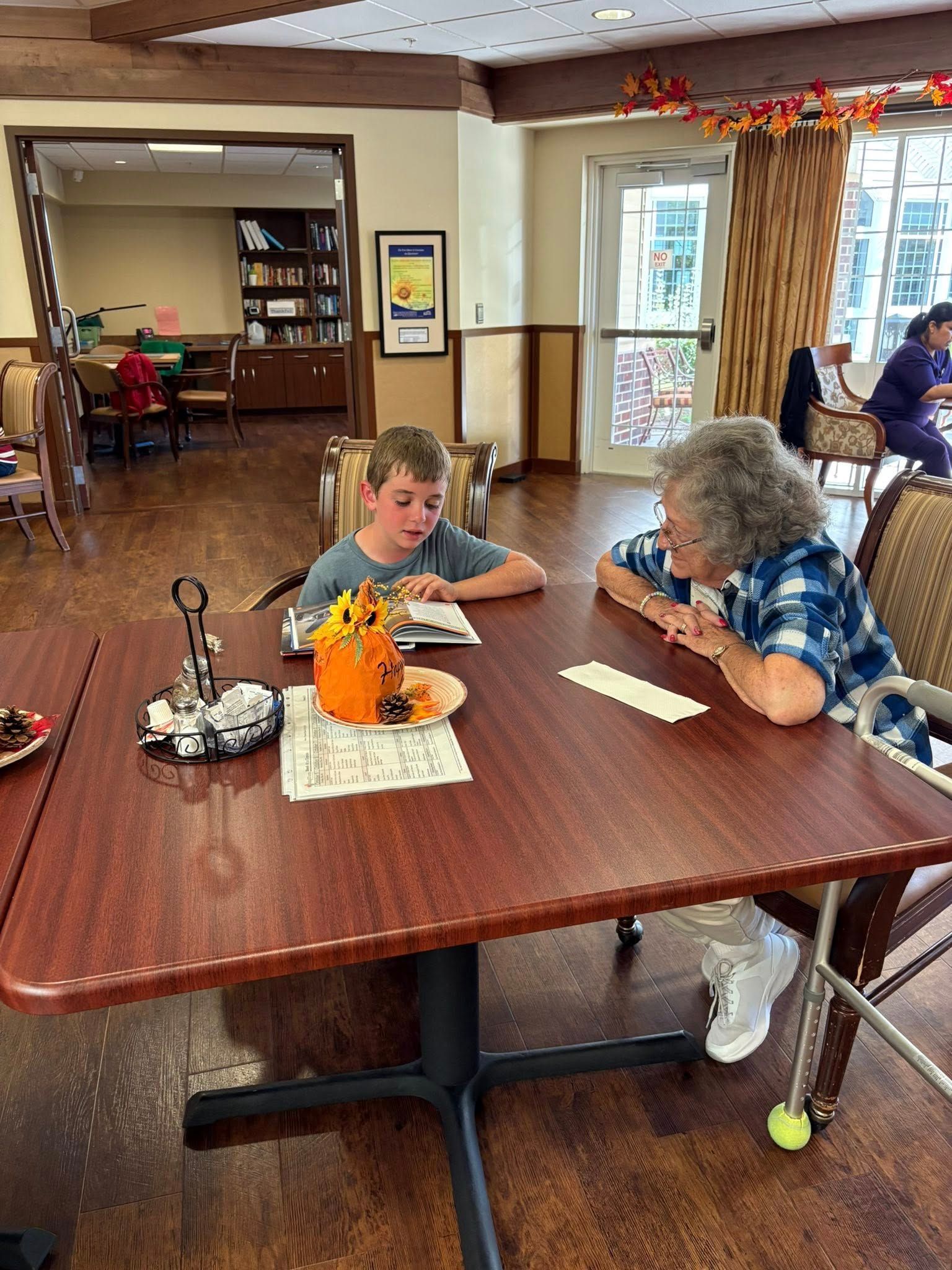 A boy and older woman sit at a table. They are in a dining area.