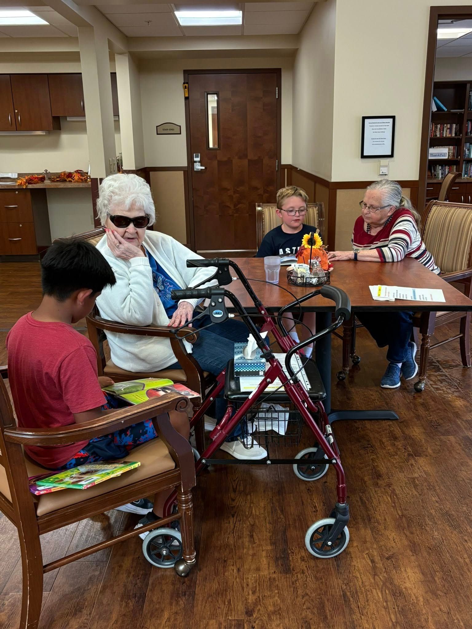 Four people at a table; one with a walker, another with dark glasses. Indoors, library or community room.