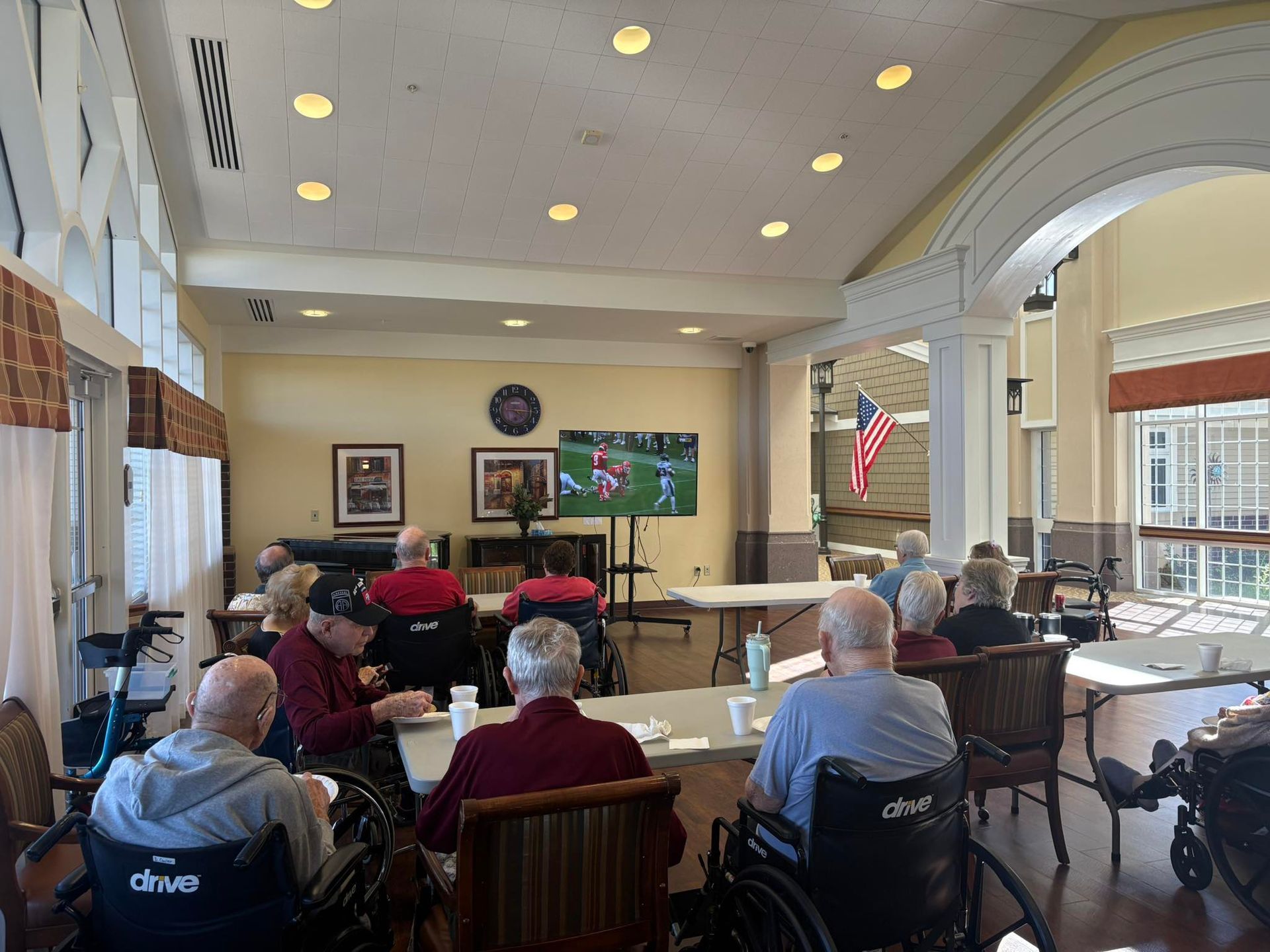 Seniors watch TV in a bright room. Some are in wheelchairs, tables in front of them. American flag visible.