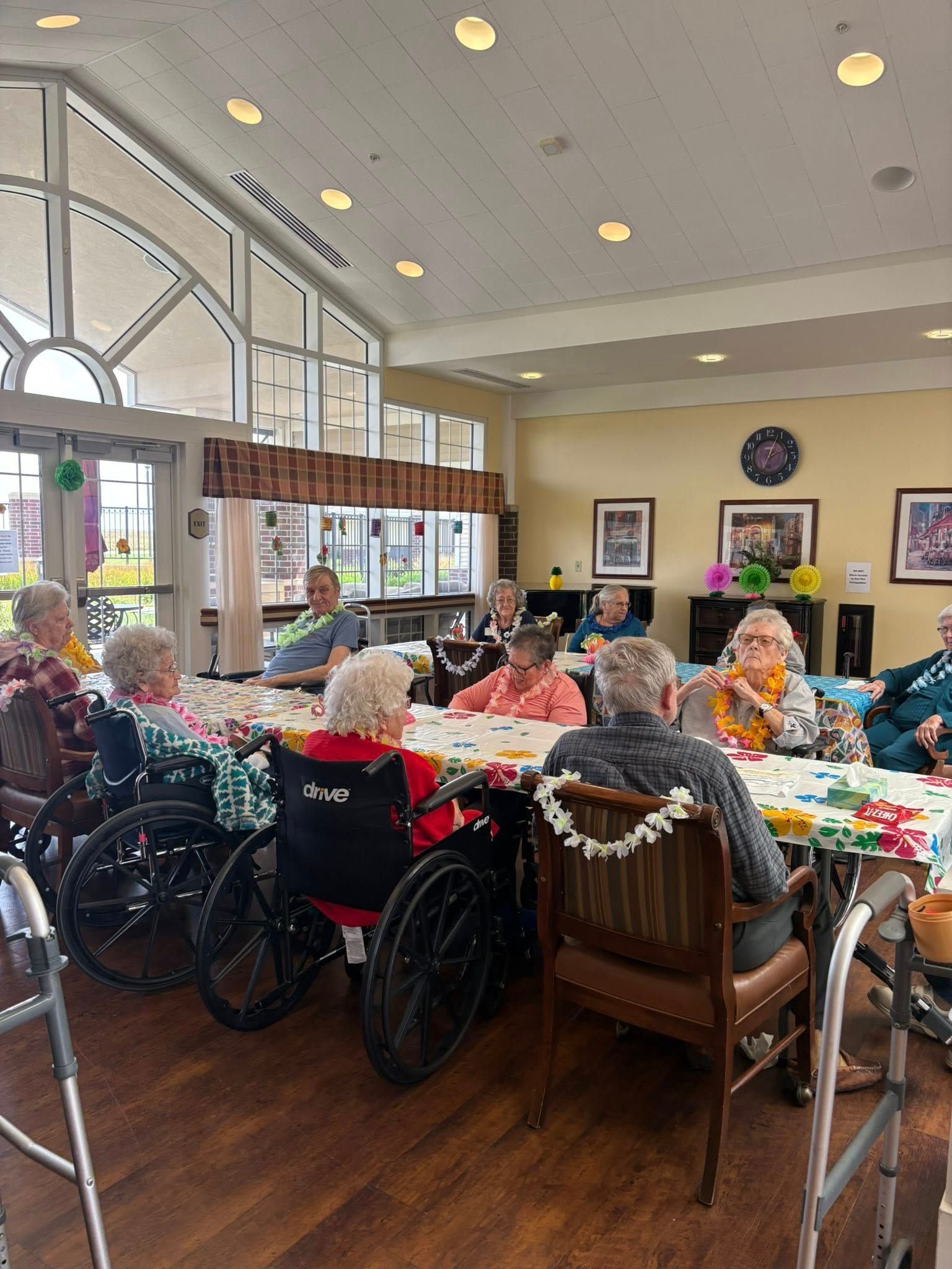 Seniors in a brightly lit room gather around a table, possibly playing a game or crafting. Some use wheelchairs.