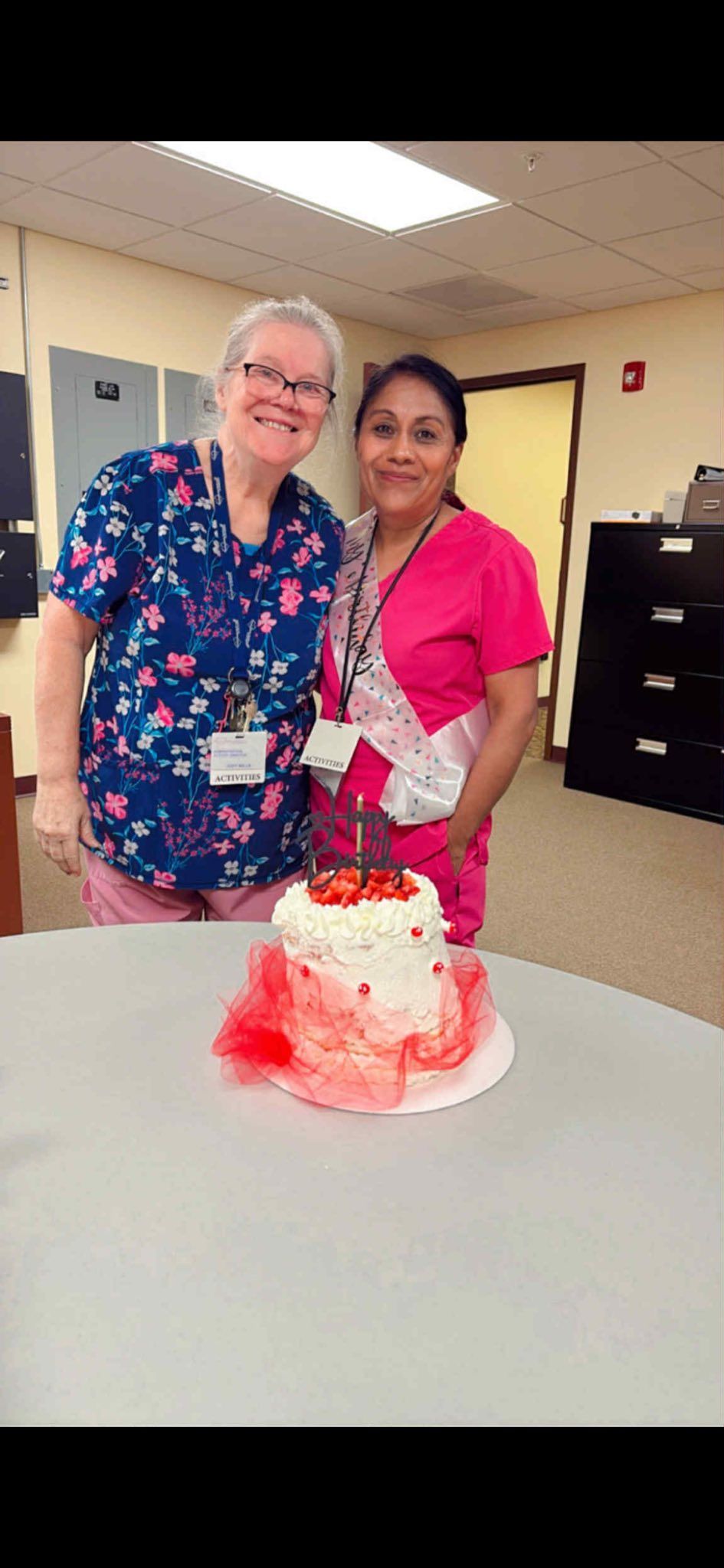 Two women standing by a cake. One is wearing a blue floral shirt and glasses. The other is wearing a pink shirt and a sash.