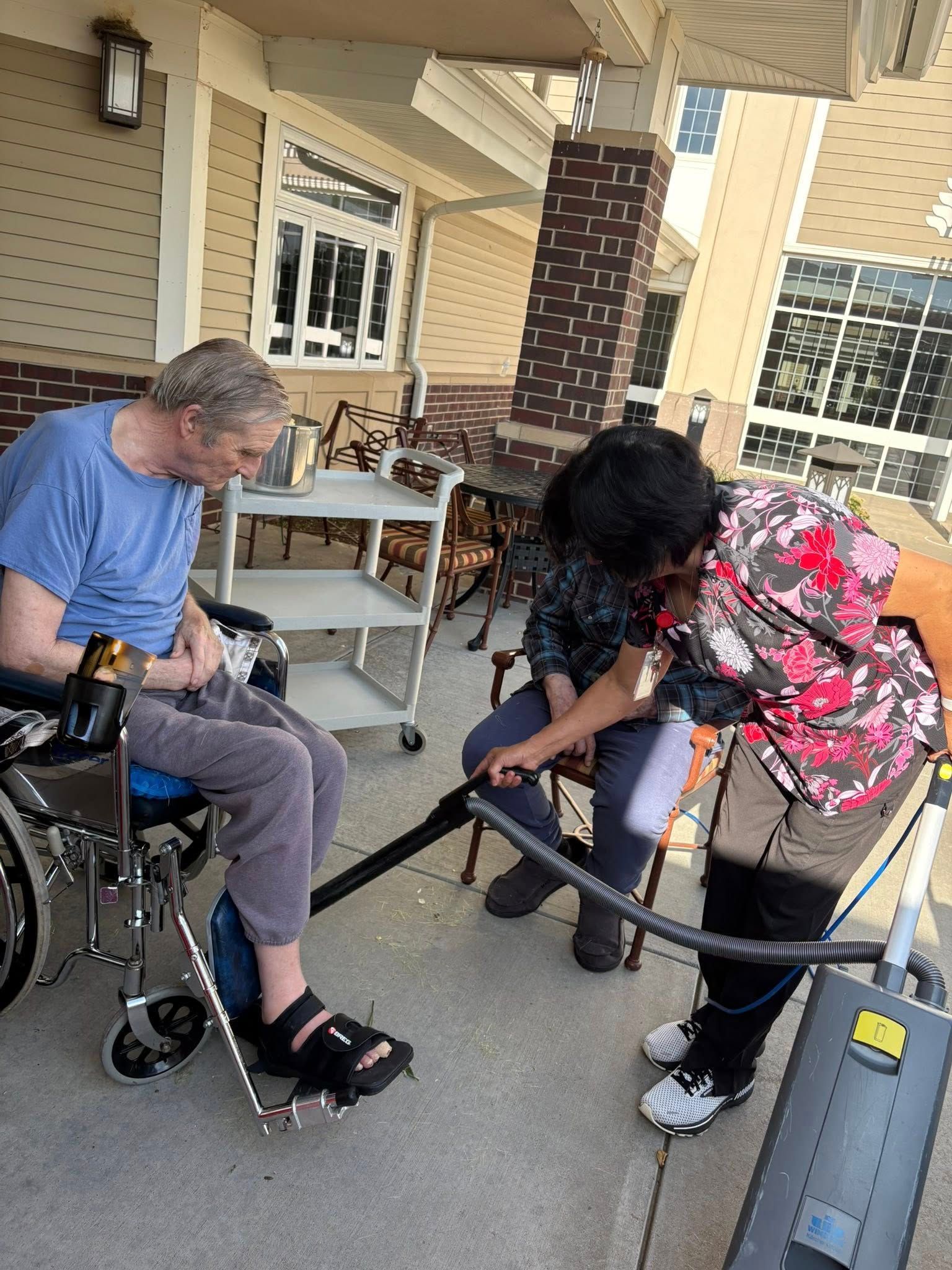 Man in wheelchair assisted by a person using a vacuum outdoors.