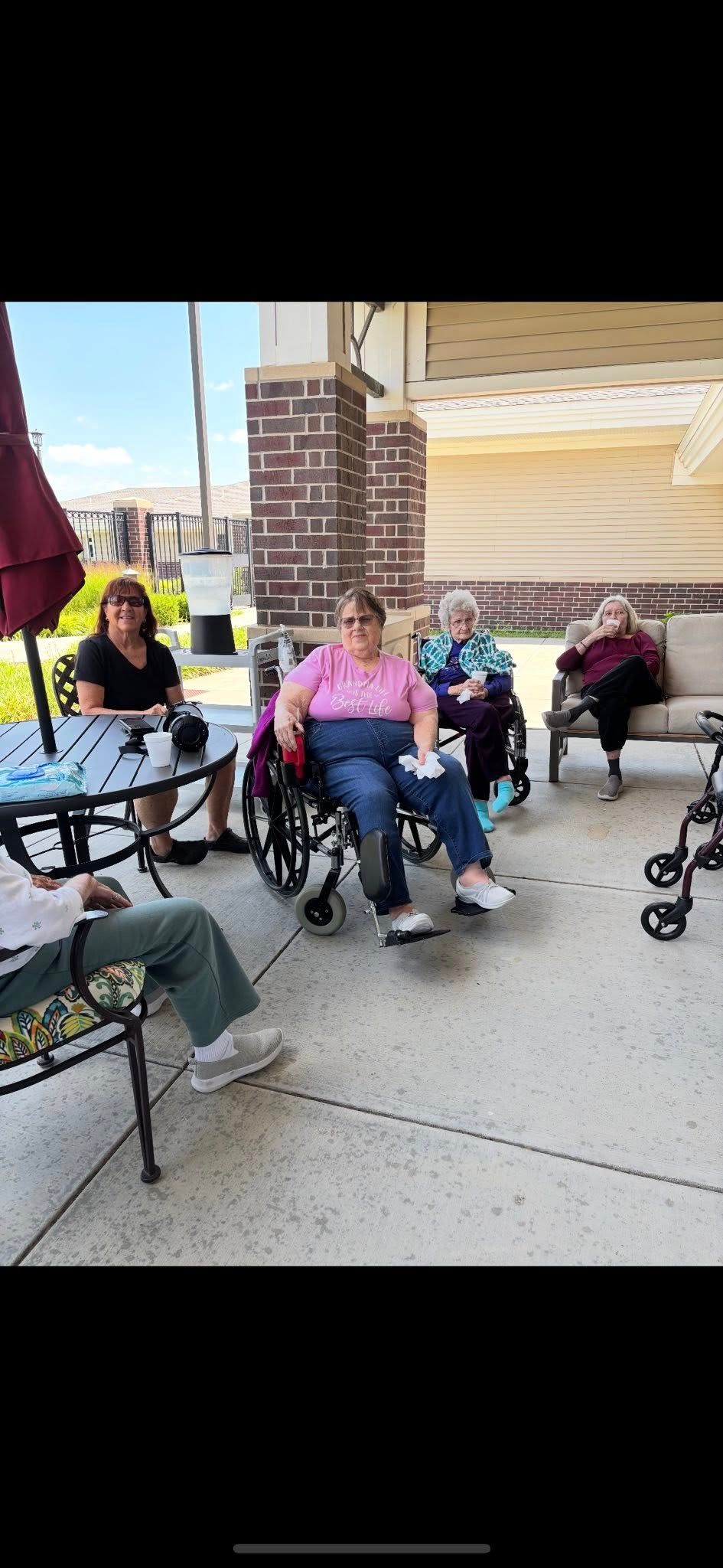 Group of people sitting and in wheelchairs on a patio near a building with red and brown columns.