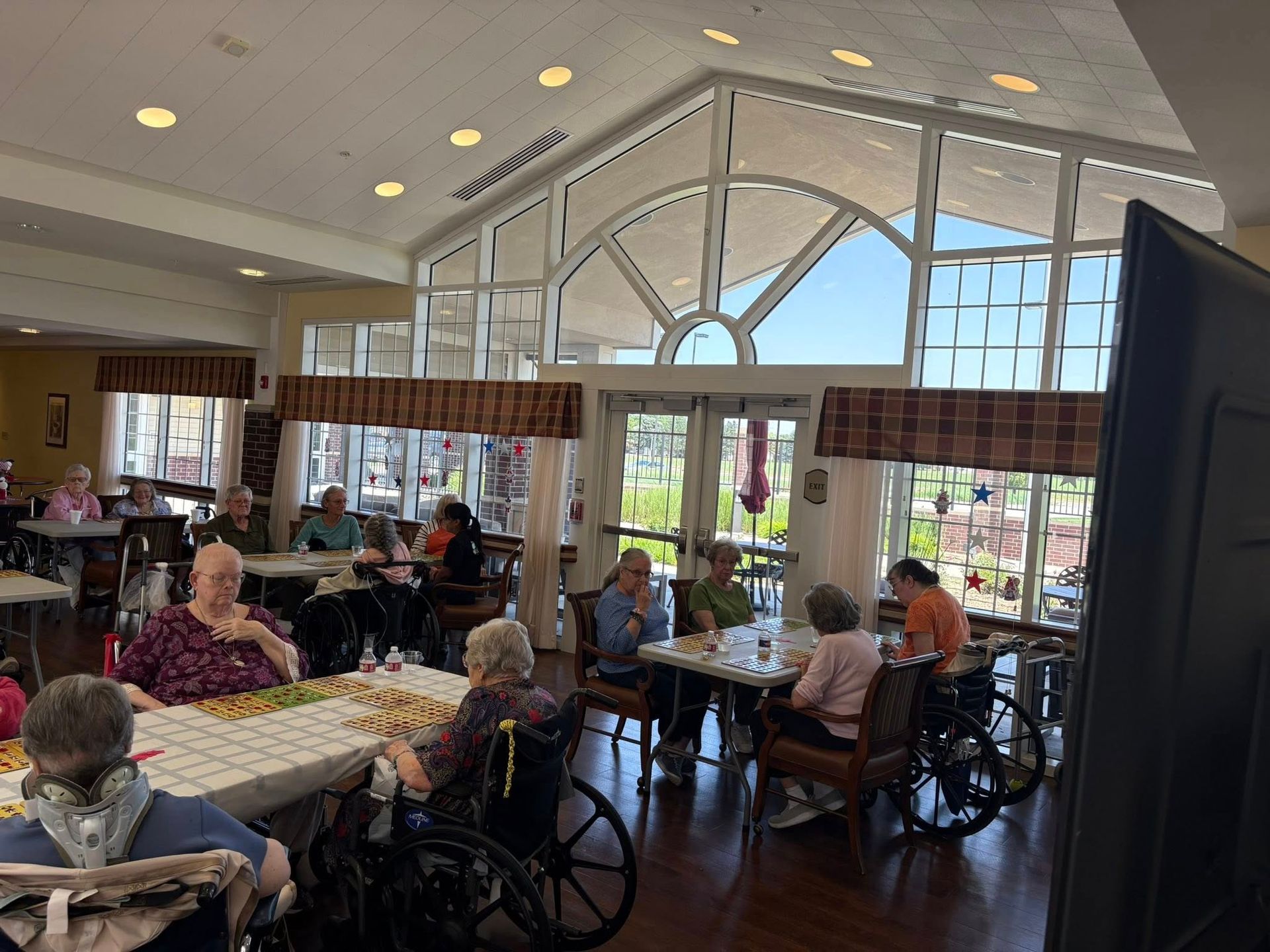 People in a light-filled dining room, some in wheelchairs, at tables. Large windows overlook an outdoor area.