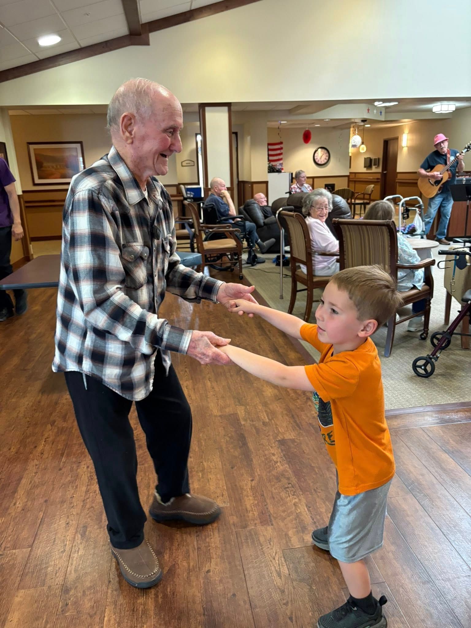 An elderly man and a young boy dance, holding hands, in a room with other people.