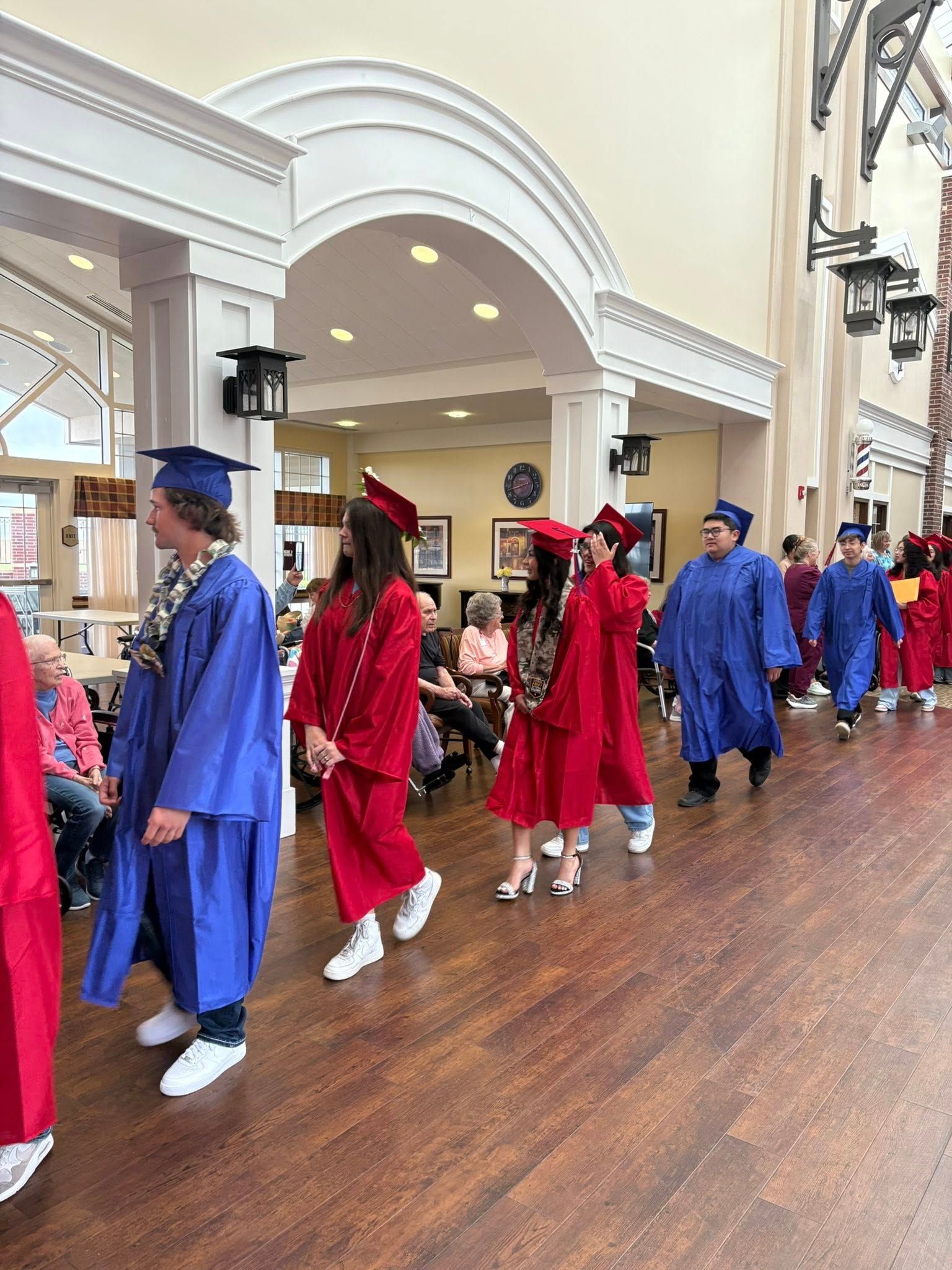 Graduates in red and blue robes with caps, walking down a hallway; some wearing leis.