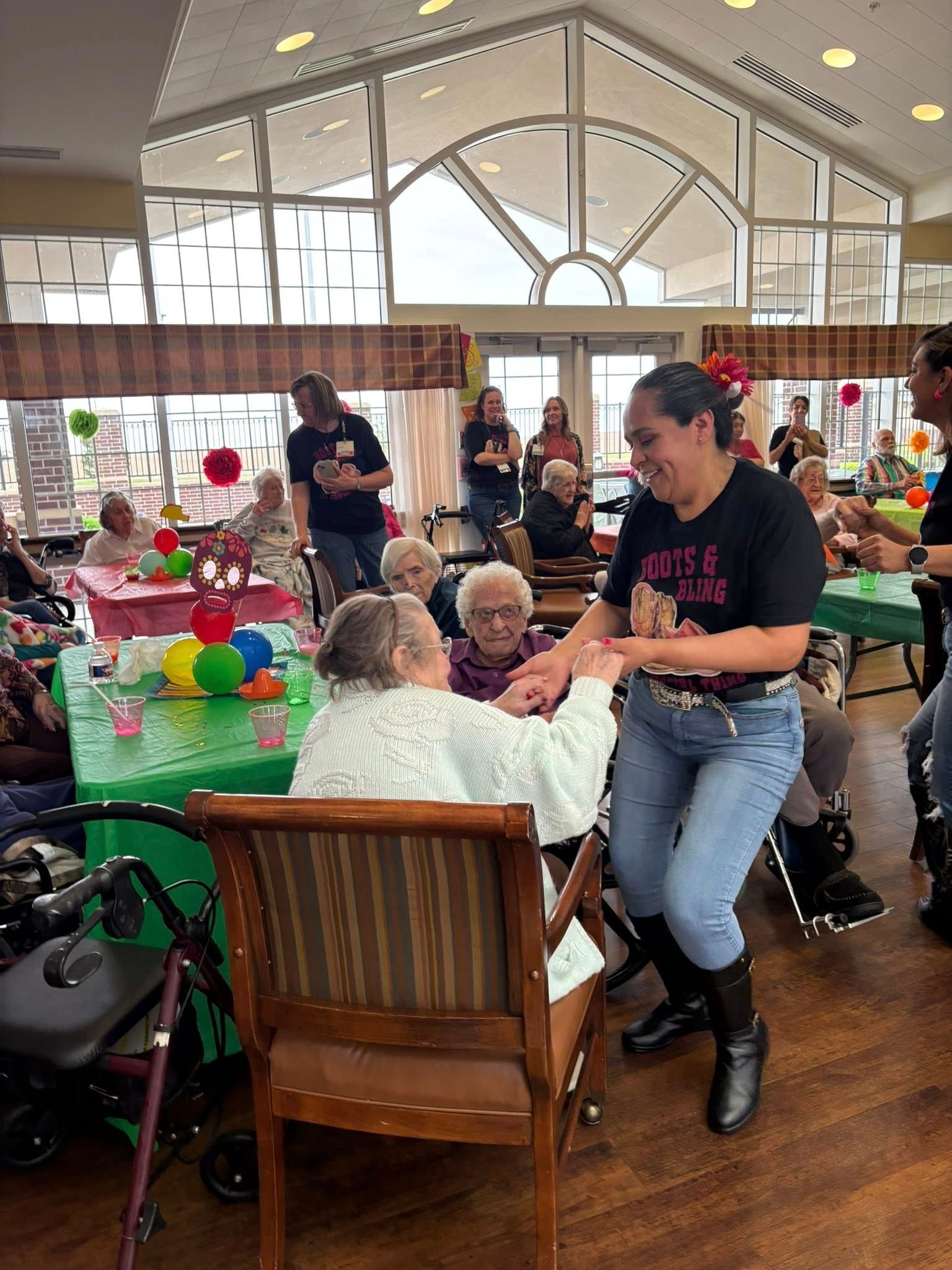 Woman dancing with seated elderly person at a party, other people watch. Bright room with decorations.