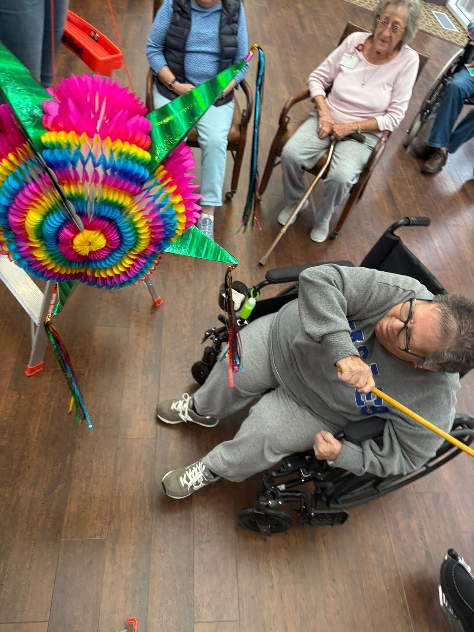Person in wheelchair swinging at a colorful pinata; other people watch in a room.