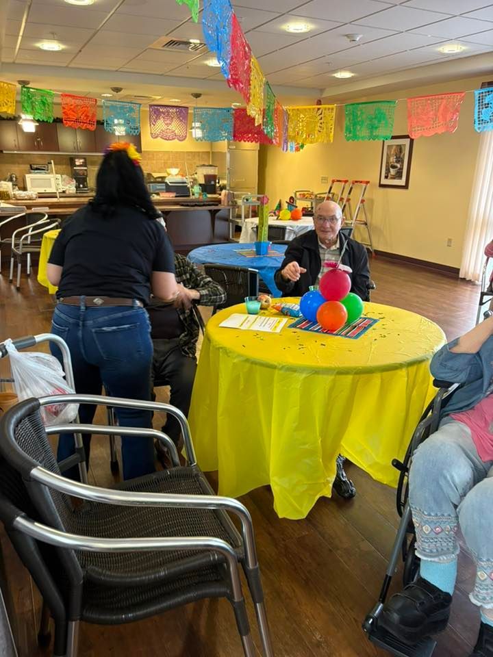Fiesta-themed event: people in a room decorated with colorful banners, a man sits at a yellow table with balloons.