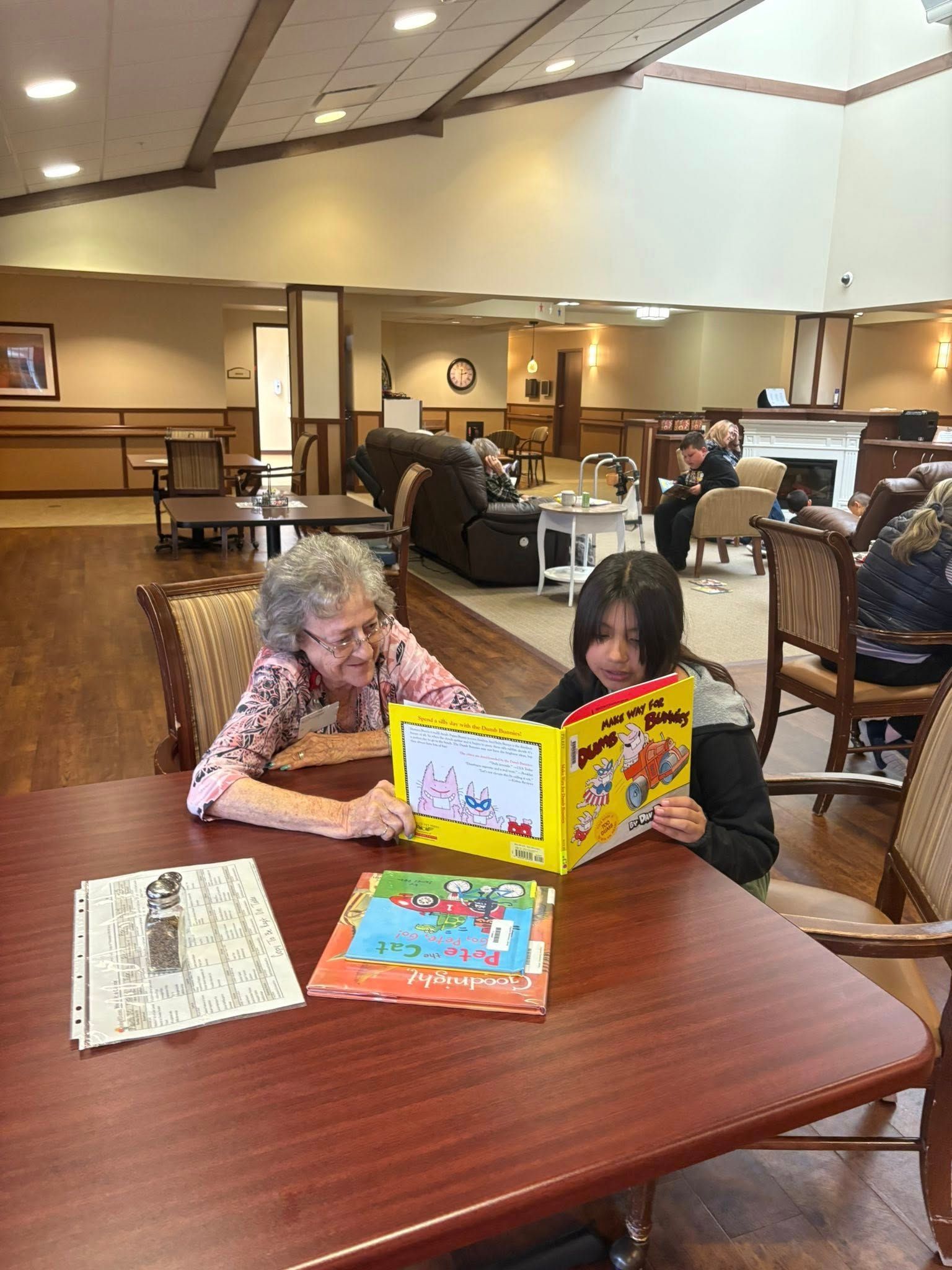 An older person and a child read a book together at a table in a well-lit community room.