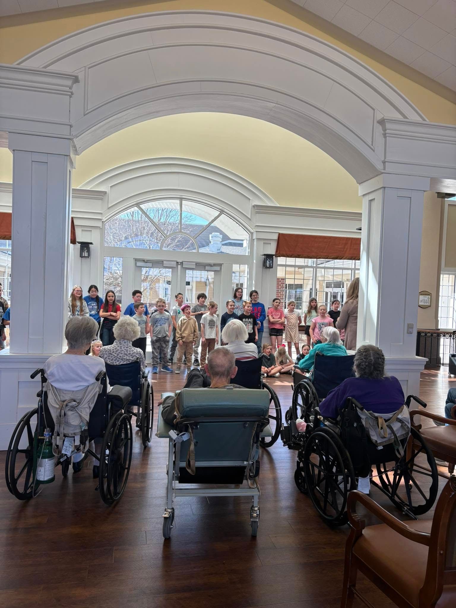 Children performing for seniors in wheelchairs under an arched hallway.