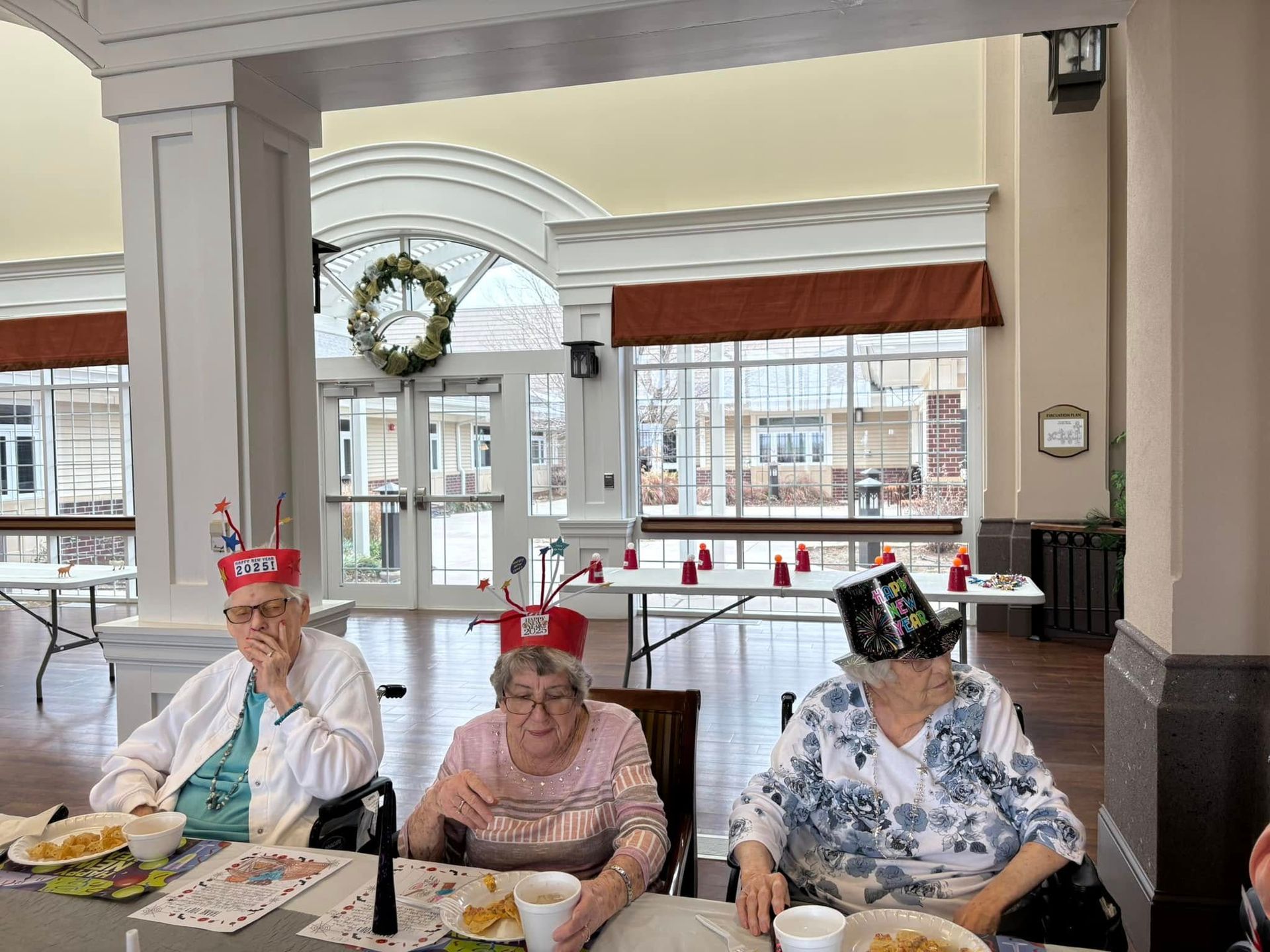 Three people wearing party hats sit at a table in a bright room with a wreath and large windows.