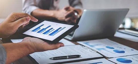 Person points at graph on a tablet, with a laptop and papers on a desk.
