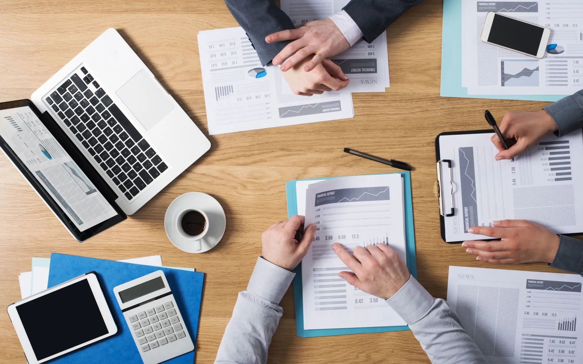 People at a table review documents, using a laptop, calculator, and tablet, with coffee and a smartphone.
