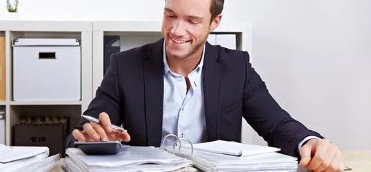 Man in suit smiling, using a calculator, papers on desk, in office setting.