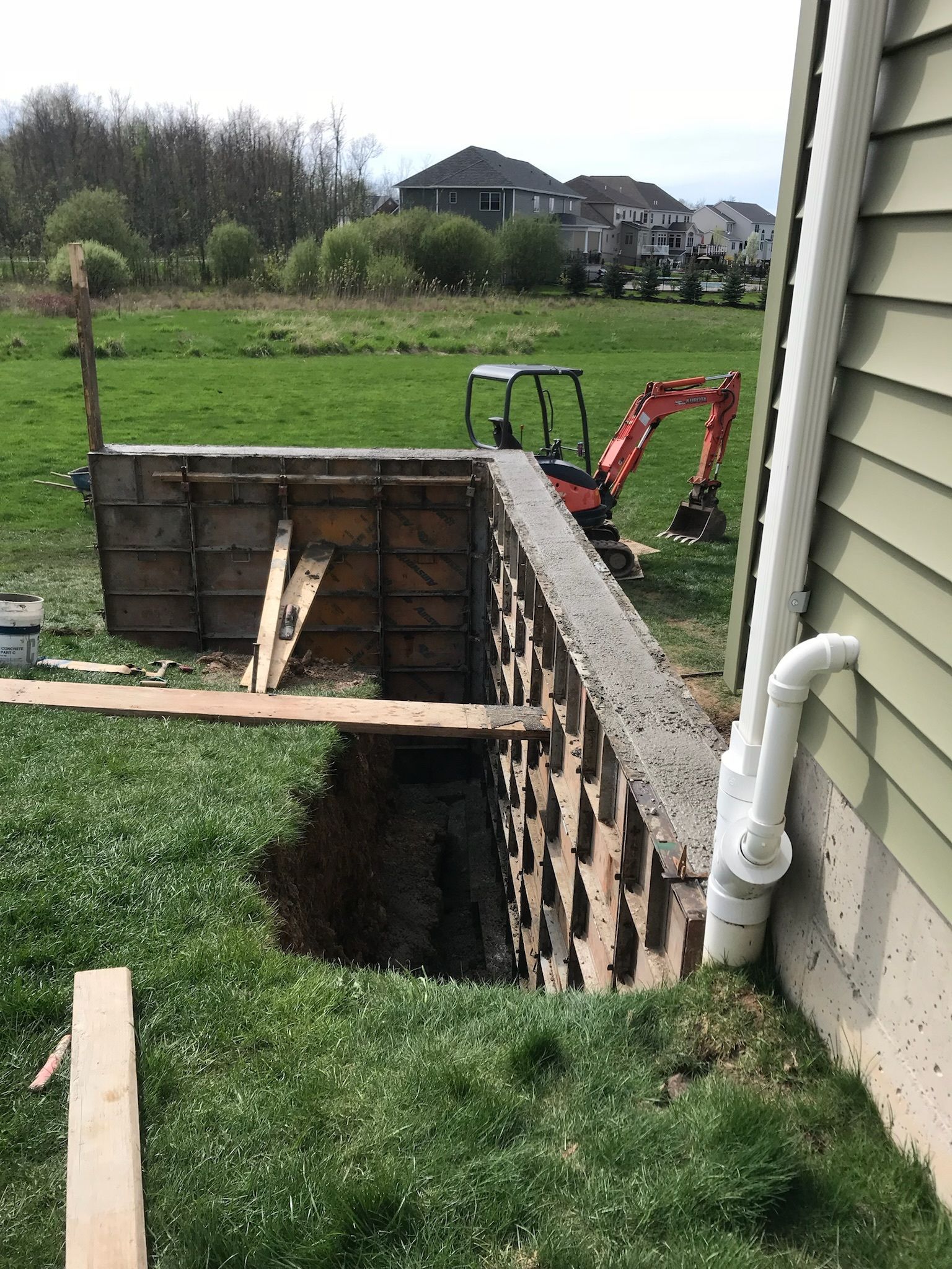 A large hole in the ground next to a house with an excavator in the background.