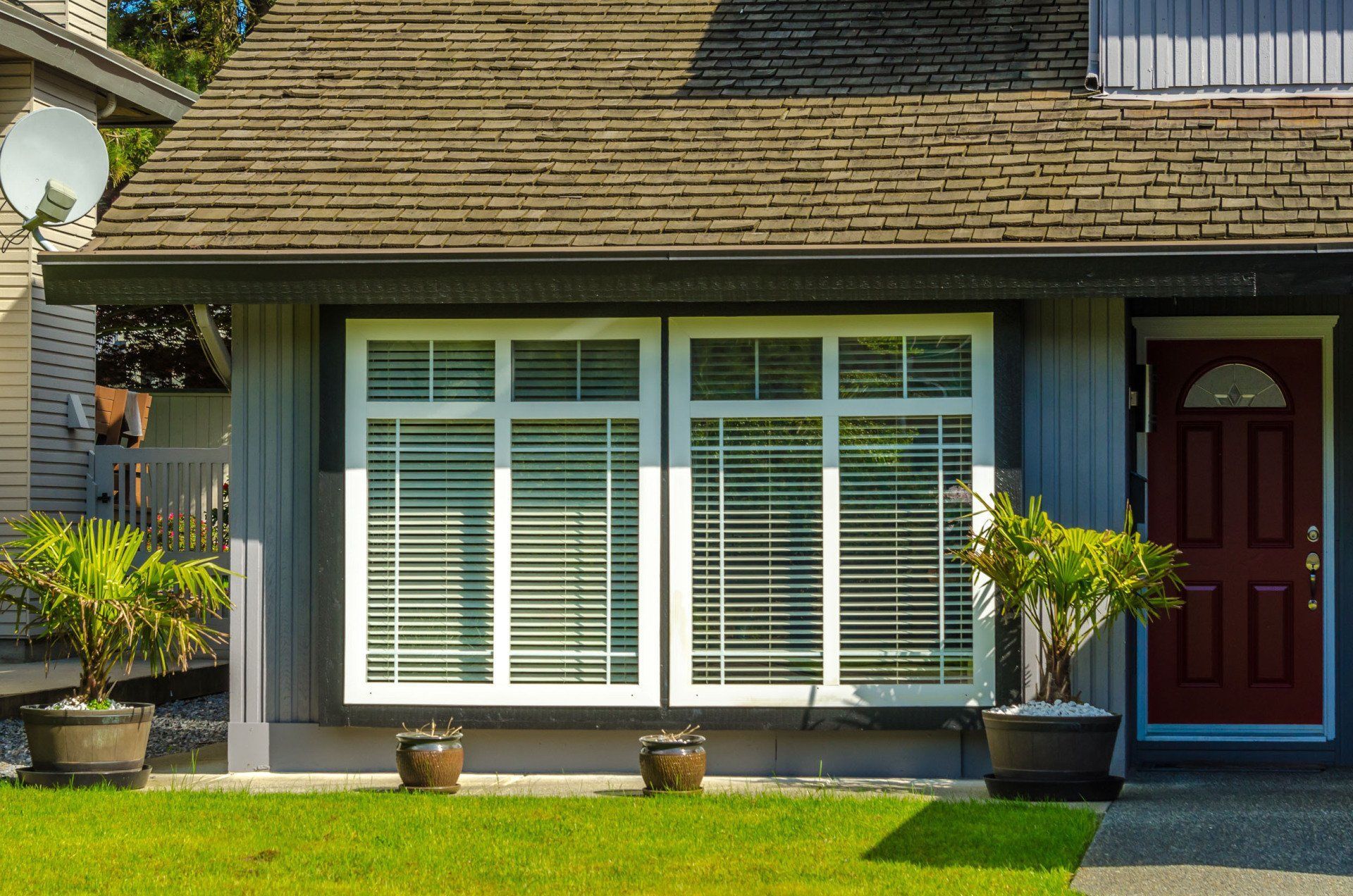 Gray house exterior with white-framed windows, red door, green lawn, potted plants, and brown roof.