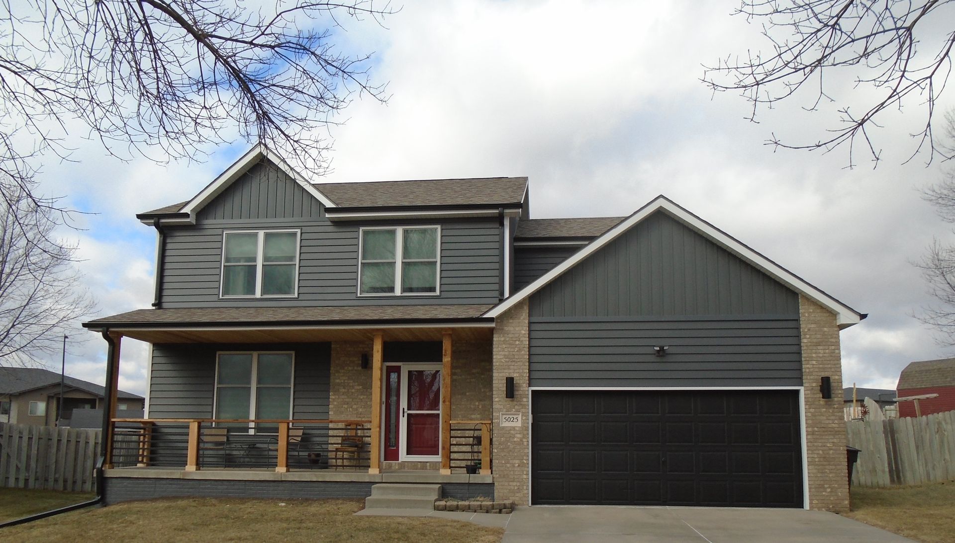 Two-story house with gray siding, brick accents, and a black garage door under a cloudy sky.