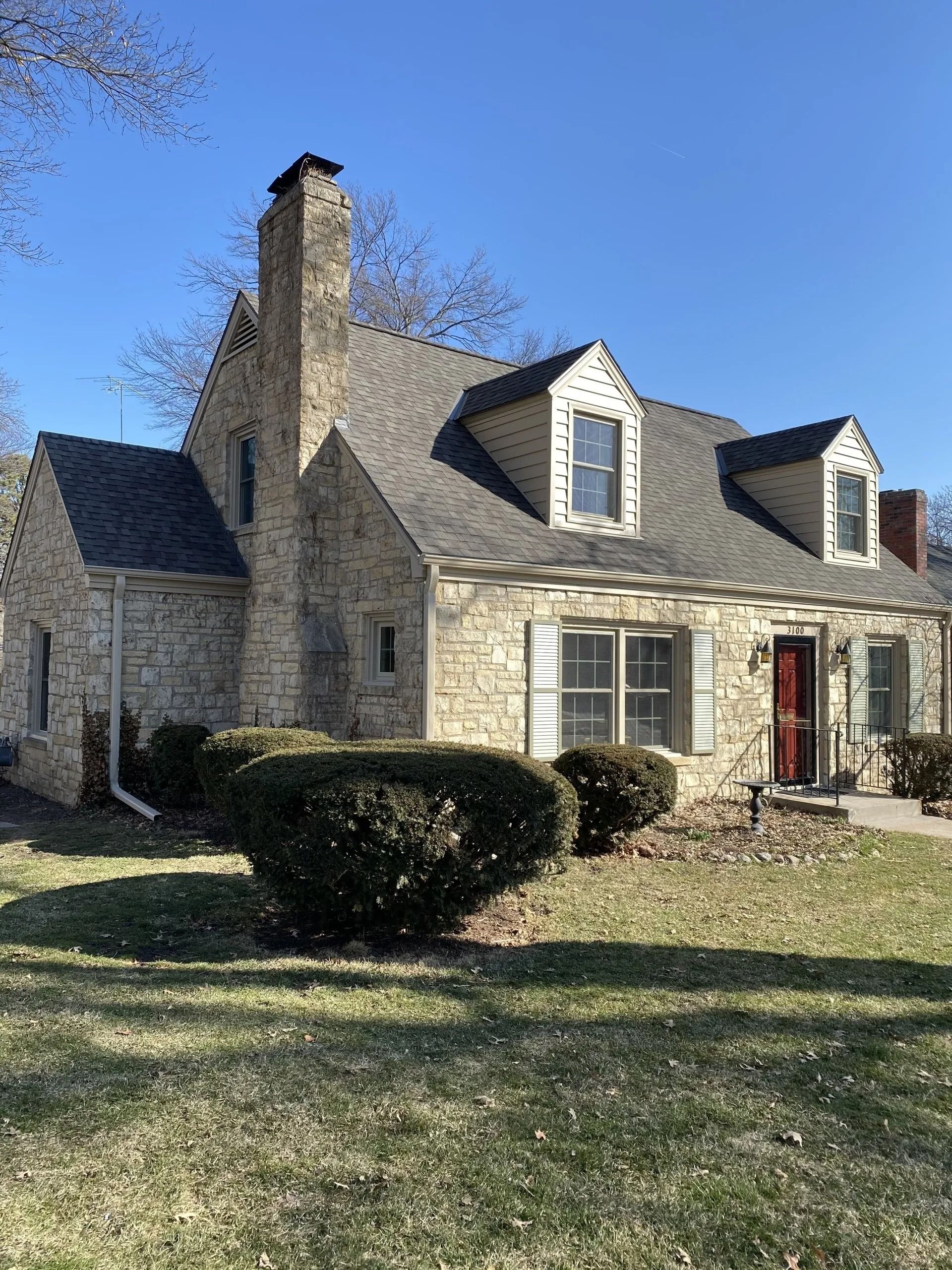 Stone-clad house with a chimney and dormers, set on a grassy lawn under a clear sky.