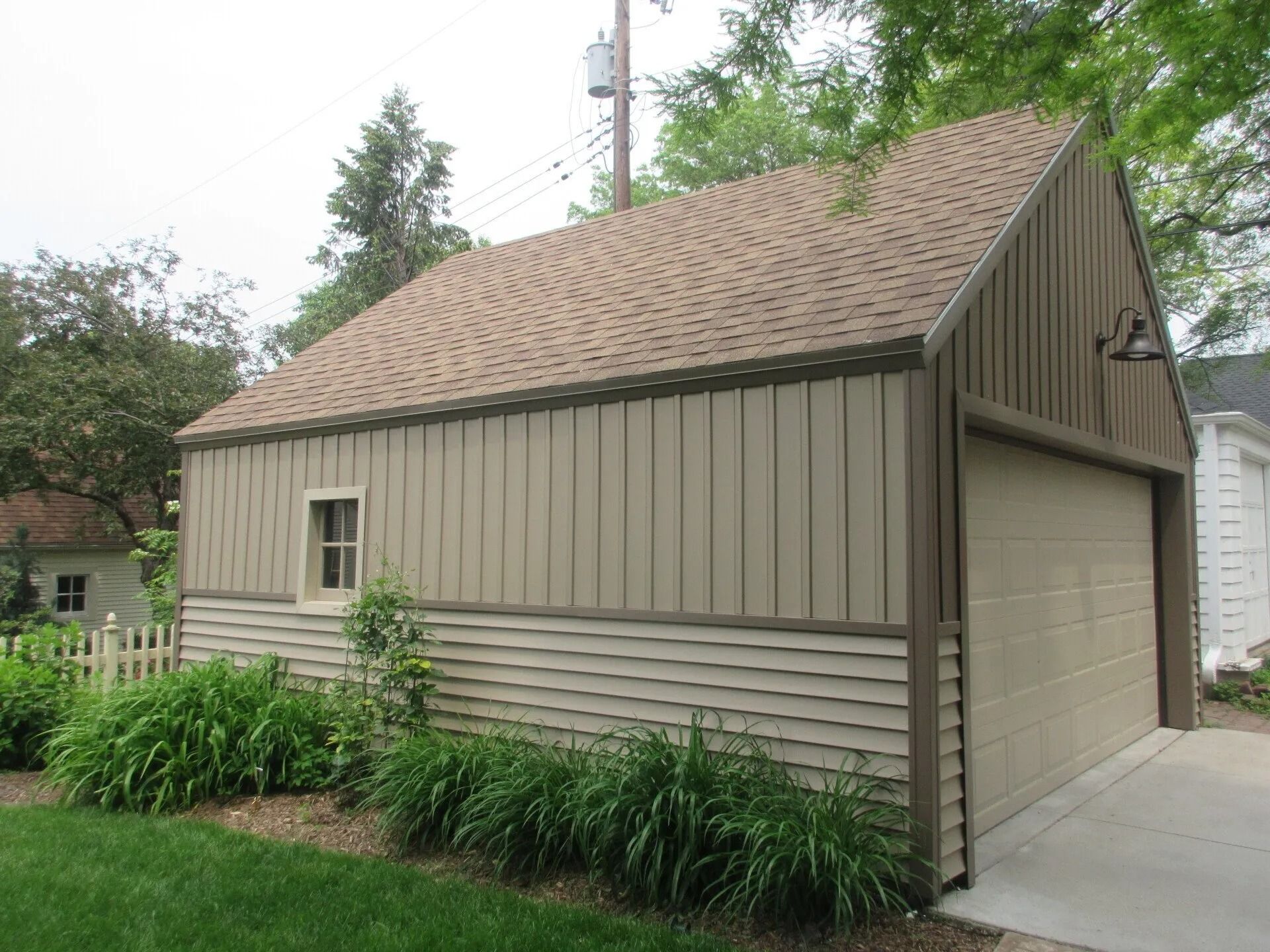 Tan garage with brown trim, roof, and a closed garage door. Window on the side, surrounded by greenery and a picket fence.