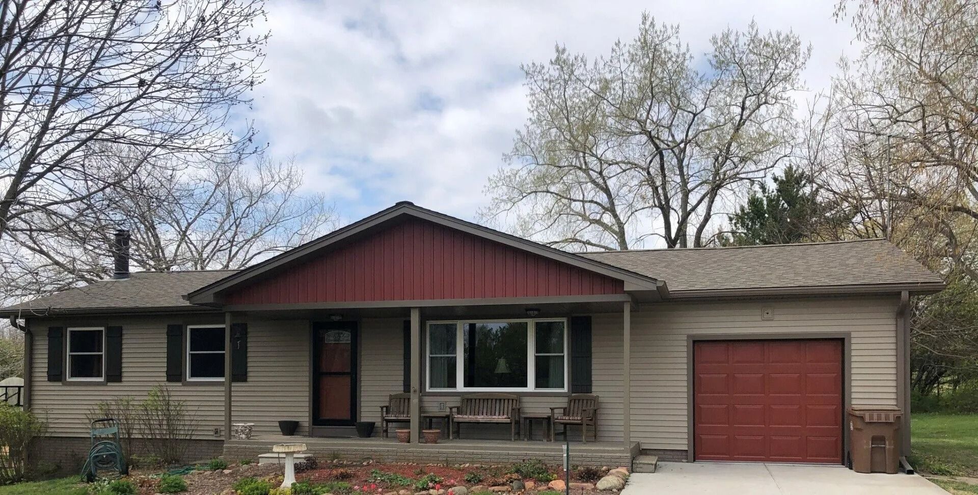 Tan house with red trim and garage door, brown roof, and trees in background.