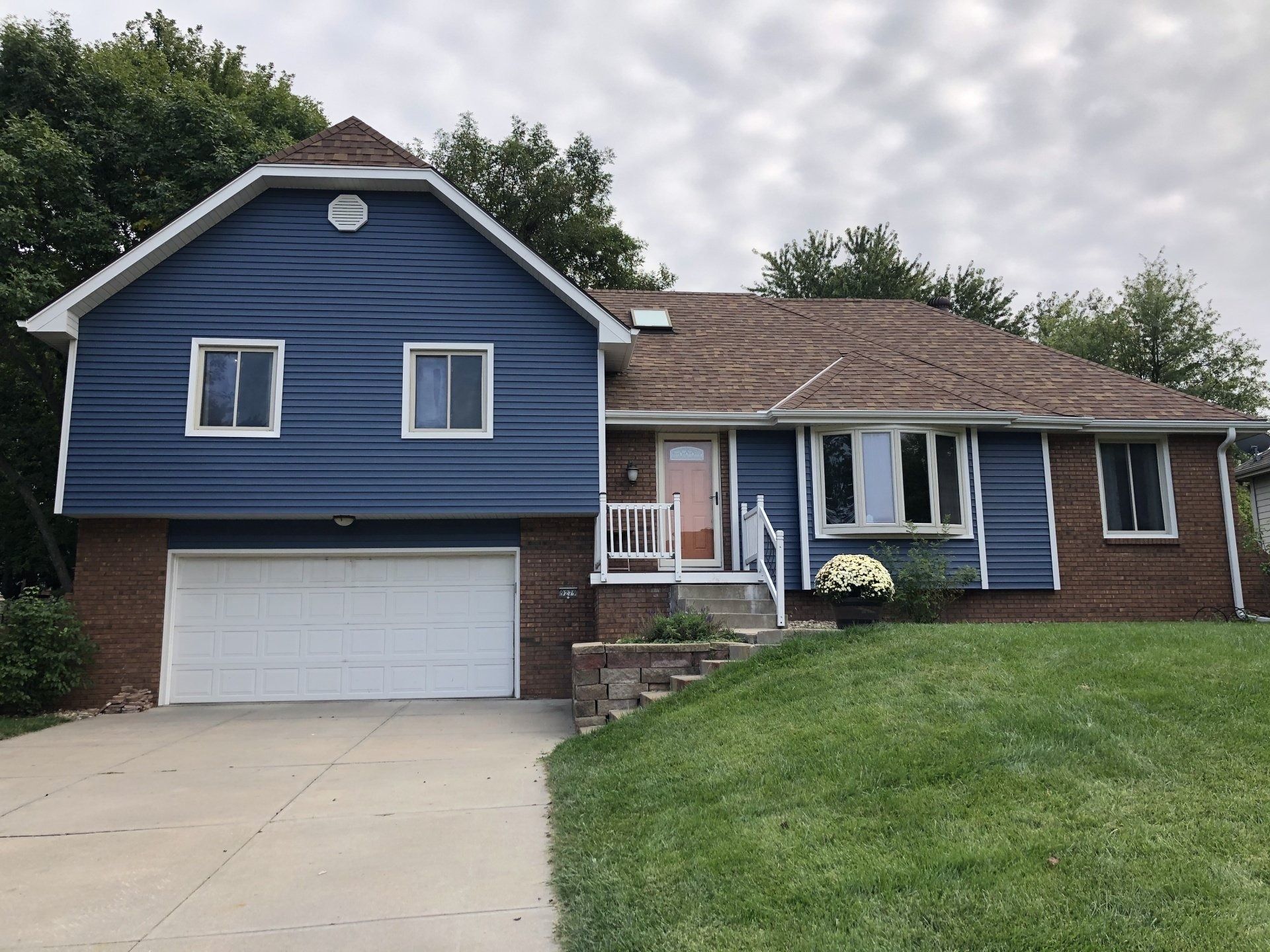 Blue-sided house with a brown roof and a white garage door, set on a green lawn with a driveway.