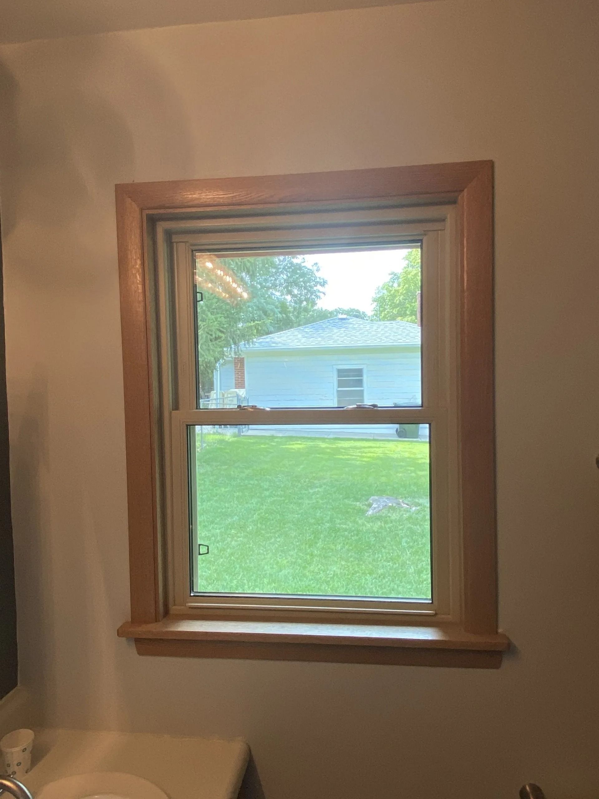 Wooden-framed window in a white-walled room, looking out onto a grassy yard and a house.