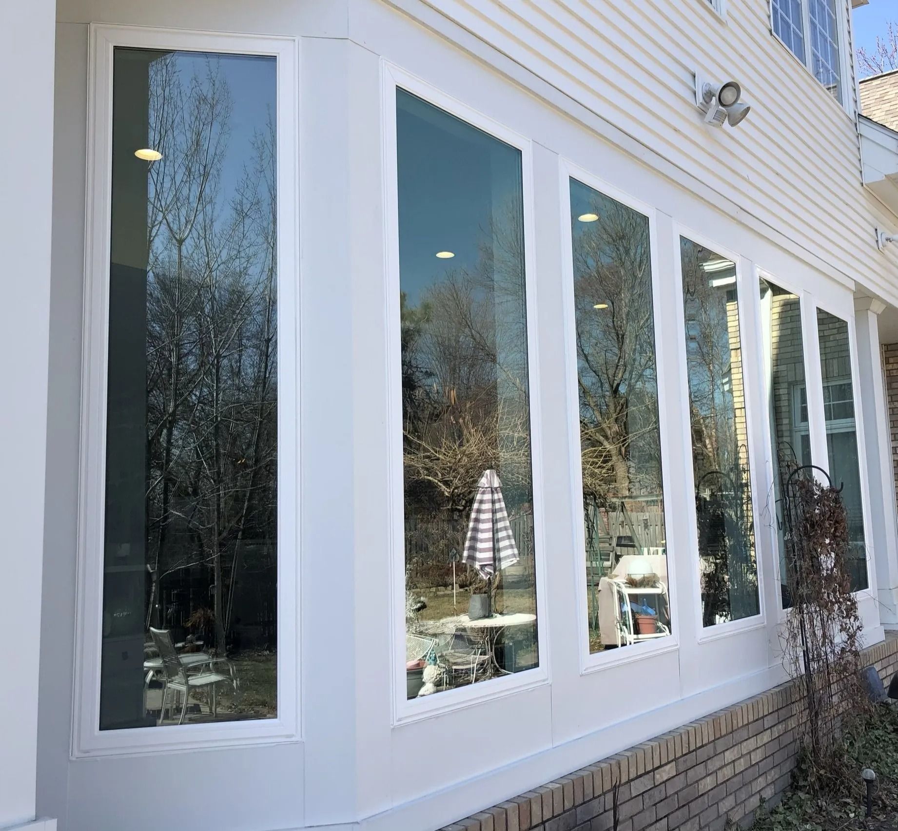 A series of tall, white-framed windows reflecting trees and a patio. The exterior is white siding and brick.