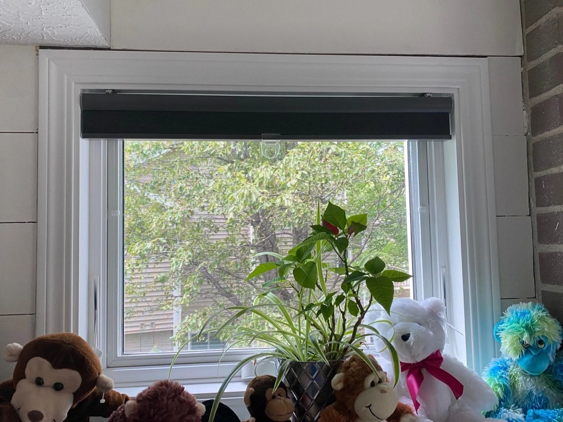 Window with a partially drawn dark shade, showcasing a green, leafy outdoor view. Plush toys sit on the windowsill.