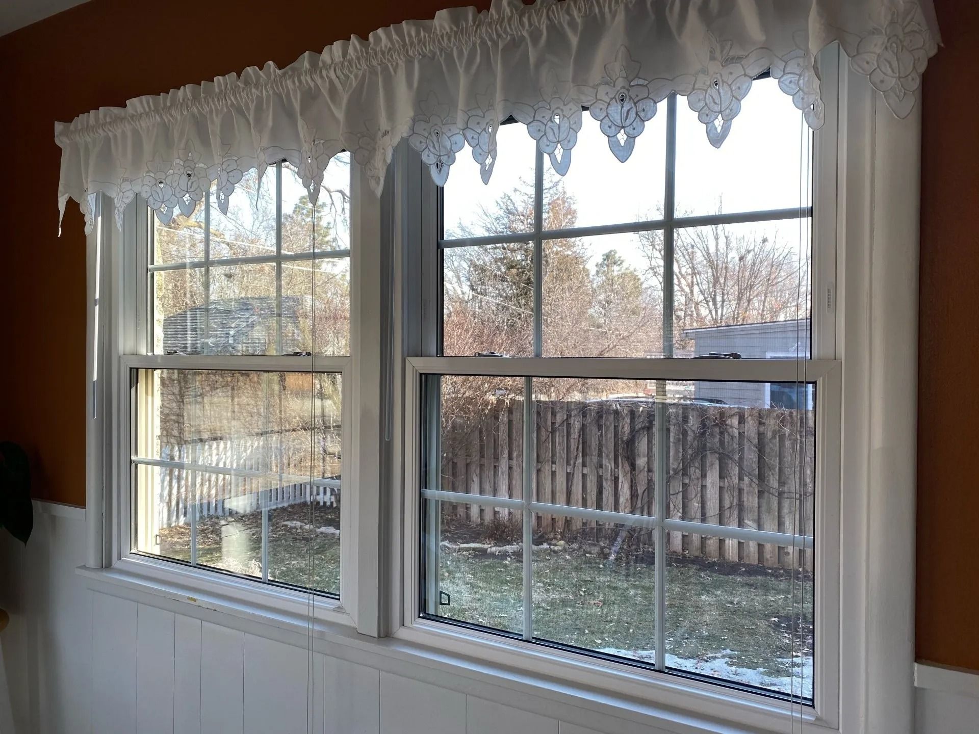 Two white-framed windows with decorative white curtains, showing a backyard with a fence.