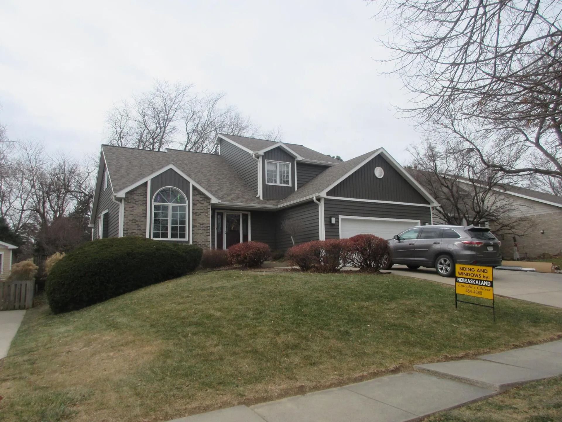 House with dark gray siding, a two-car garage, and a for sale sign on a slightly sloped lawn.
