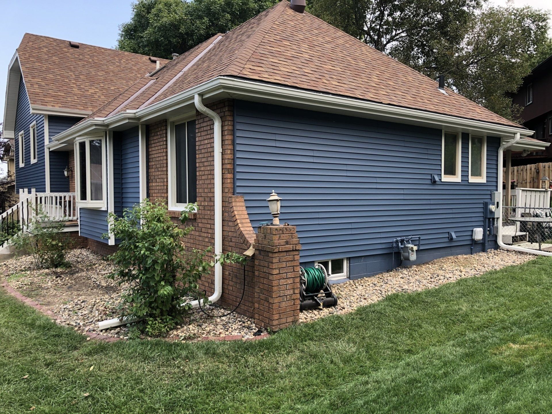 Blue house with brown roof and brick accents, surrounded by green grass and landscaping.