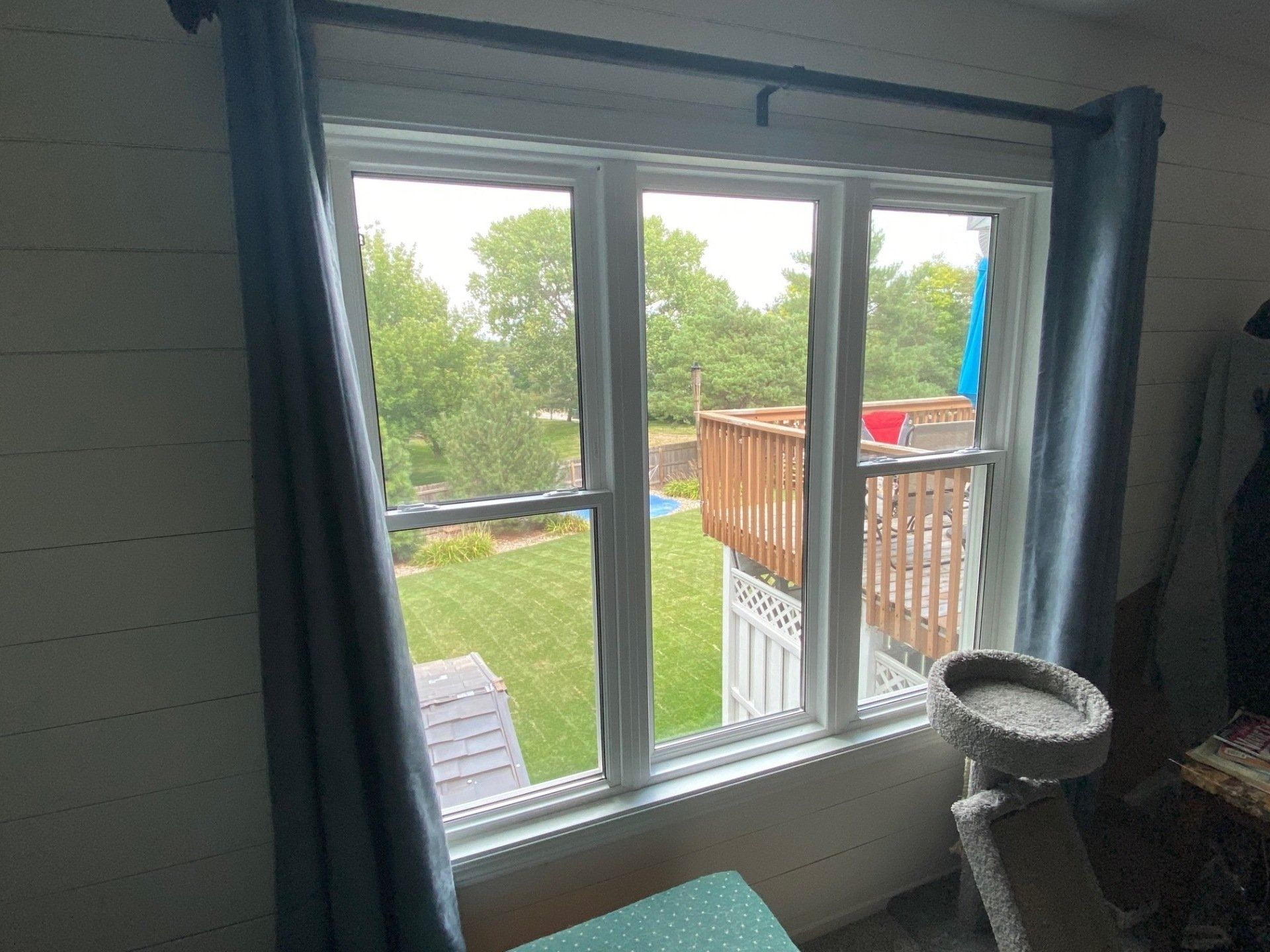 Window overlooking a backyard with green grass, a deck, and trees; blue curtains on either side.