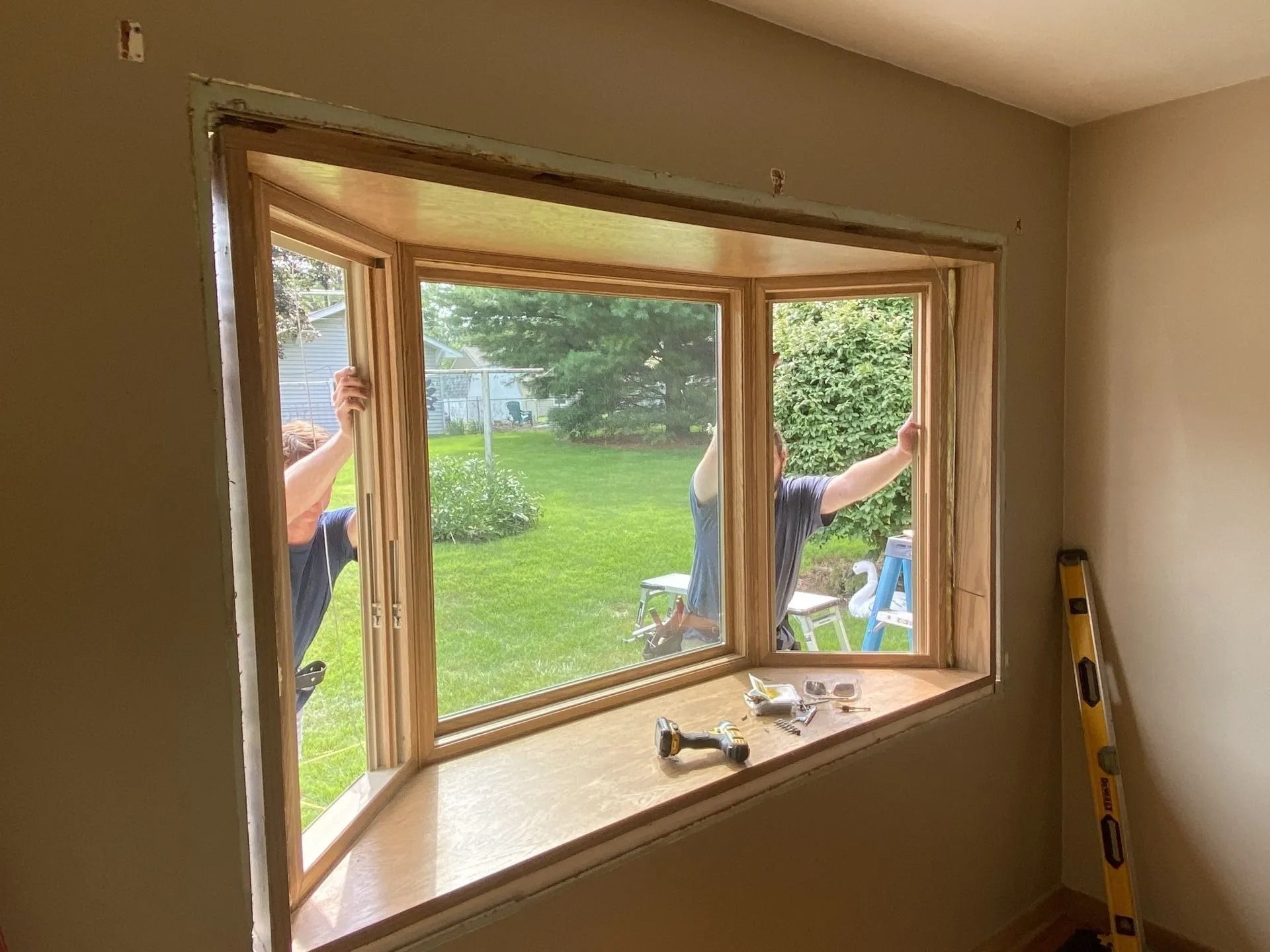 Two people installing a wooden bay window in a room with a backyard view.