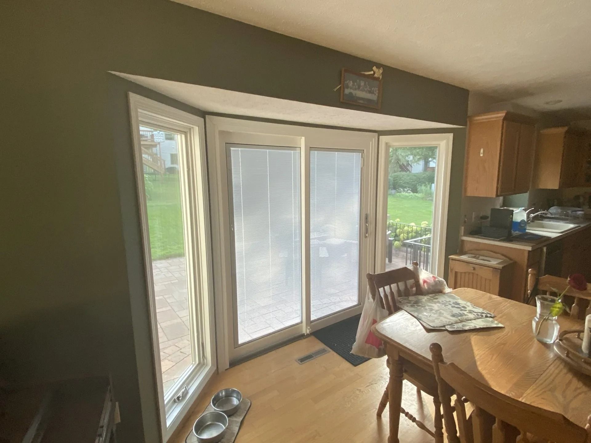 Bay window with white sliding doors and flanking windows overlooking a backyard; adjacent wood dining table and chairs.