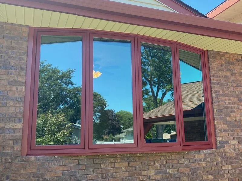 Bay window with red trim reflects trees and sky on a brick house.