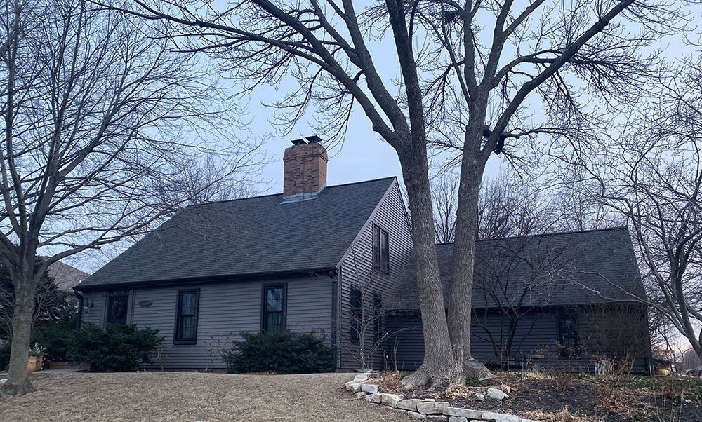 Gray house with dark roof and chimney, bare trees in front.