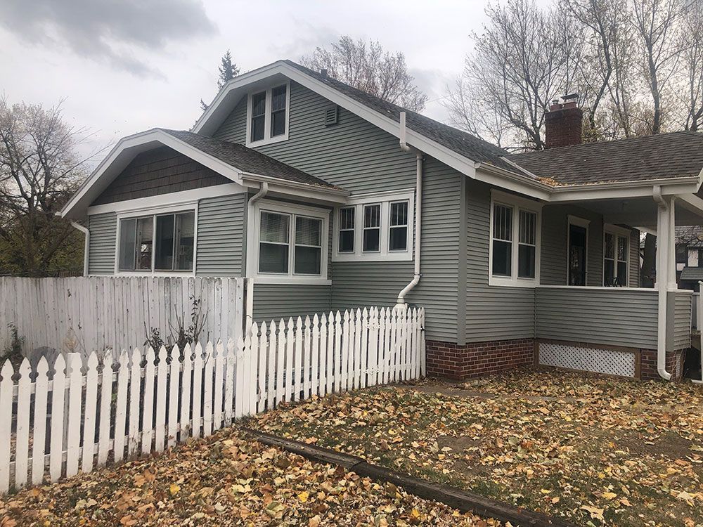 A gray house with white trim and a picket fence in front. The yard is covered in fallen leaves.
