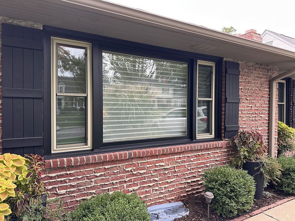 Black-framed window on a red brick house with black shutters, and tan trim. Green shrubs and plants are in front.