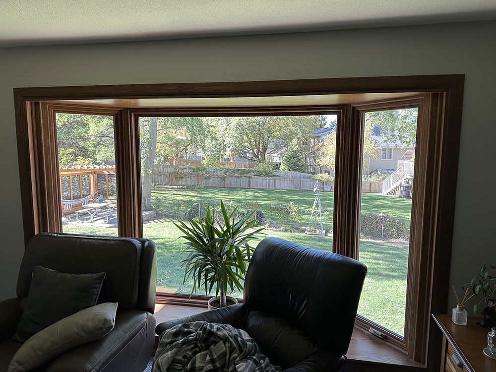 Bay window with wooden trim overlooking a backyard; two armchairs inside, plant on windowsill.