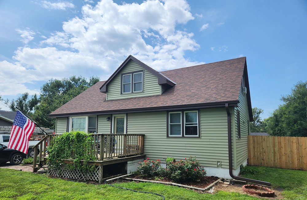 Green house with brown roof, wooden deck, American flag on the left, blue sky with clouds.