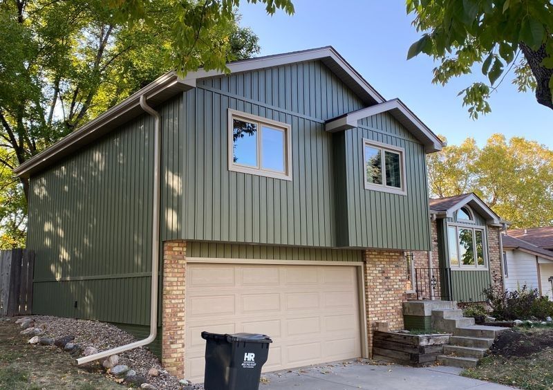 Green-sided two-story house with beige garage door, brick and tan trim. Trees in background, sunny day.