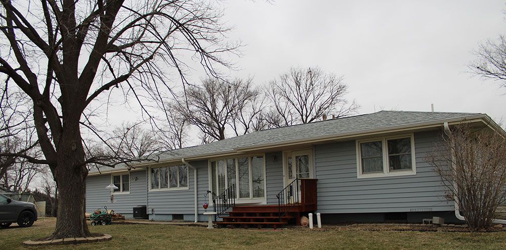 Blue-sided ranch-style house with brown porch and bare trees in yard, cloudy sky.