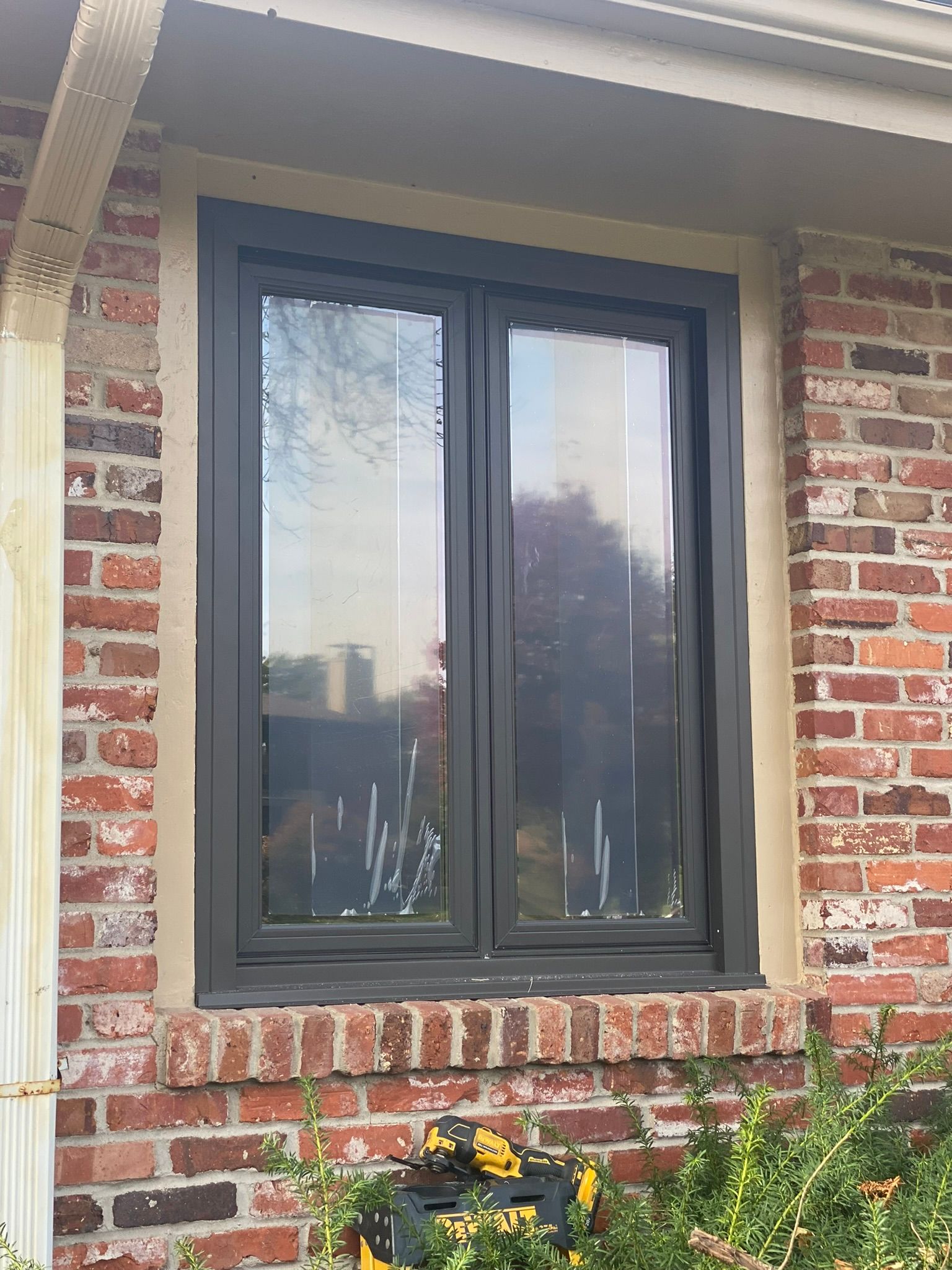 Black-framed window in a red brick wall, reflecting trees and sky, surrounded by tan trim.
